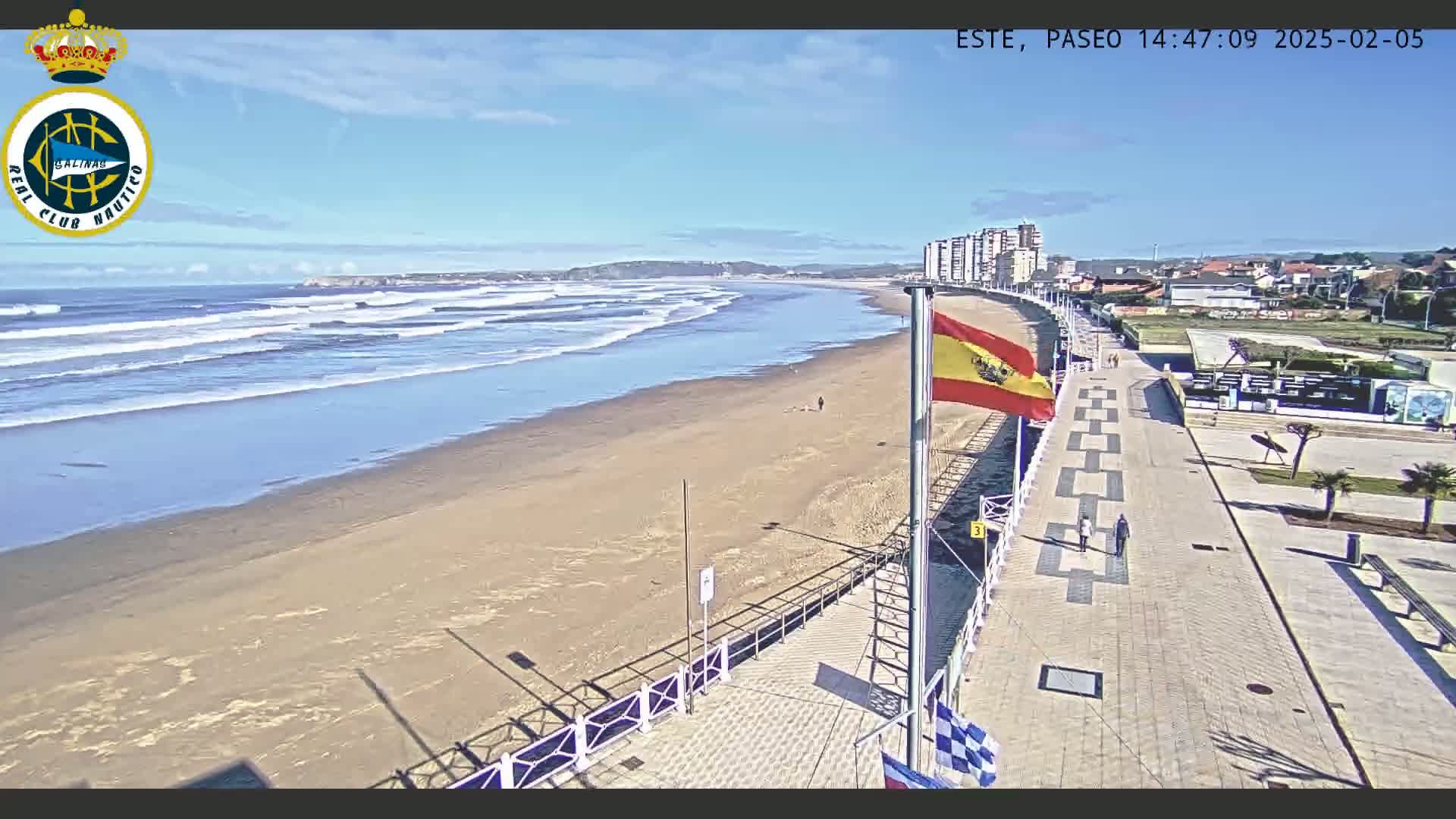 A sandy beach with gentle waves under a clear blue sky is viewed from a boardwalk with buildings in the background.