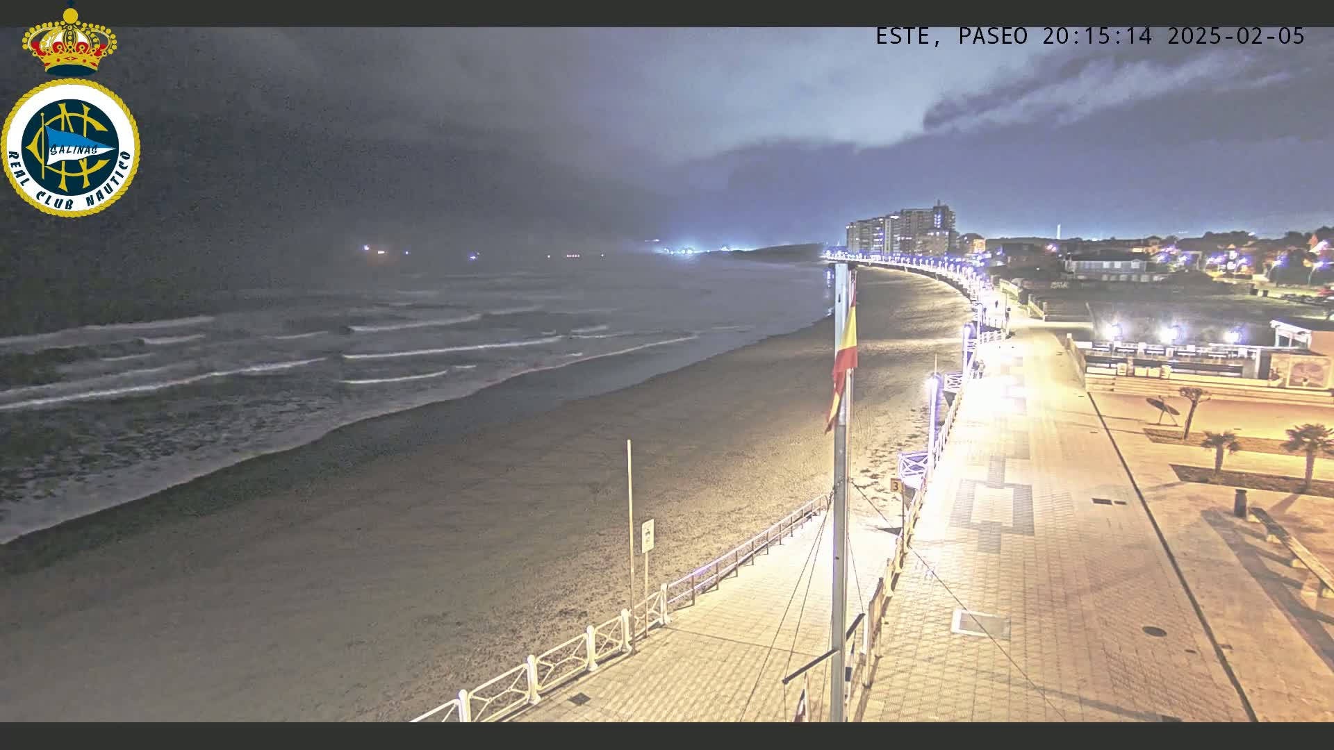 A nighttime beach scene shows gently lapping waves on a sandy shore, a boardwalk lit with streetlights, and buildings in the distance under a cloudy sky.