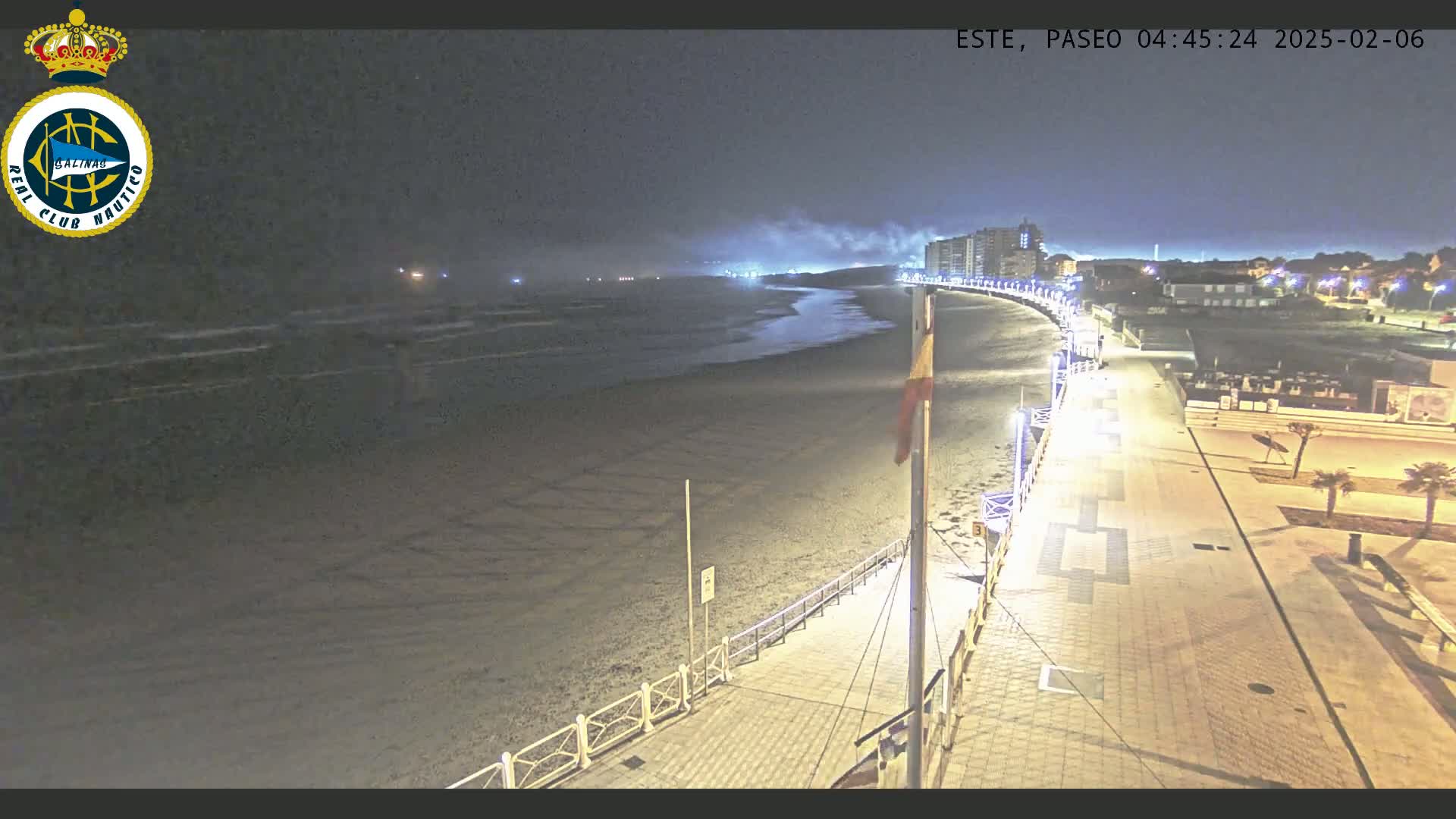 A nighttime view of a mostly empty beach with a paved walkway, some buildings in the distance, and calm waves under a hazy sky.