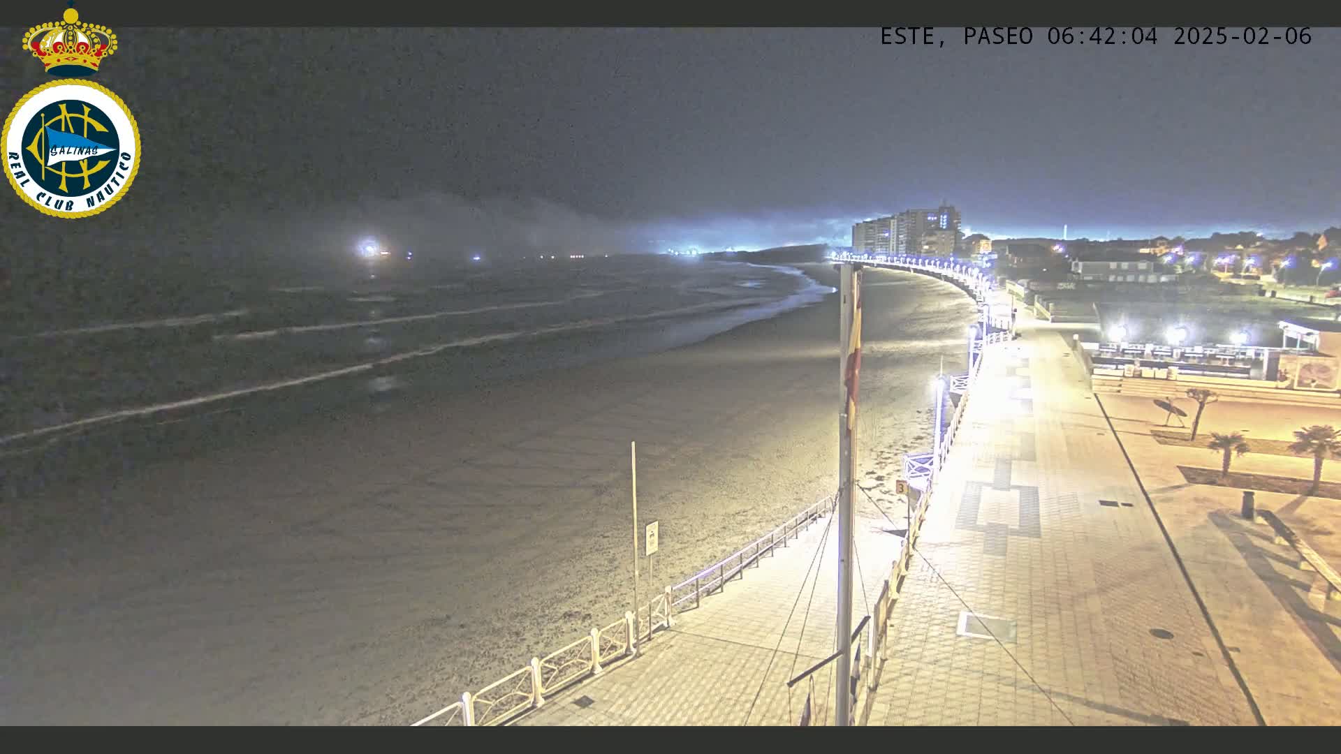 A nighttime beach scene shows a paved walkway leading to a sandy beach with gentle waves under a hazy sky, and buildings in the distance.