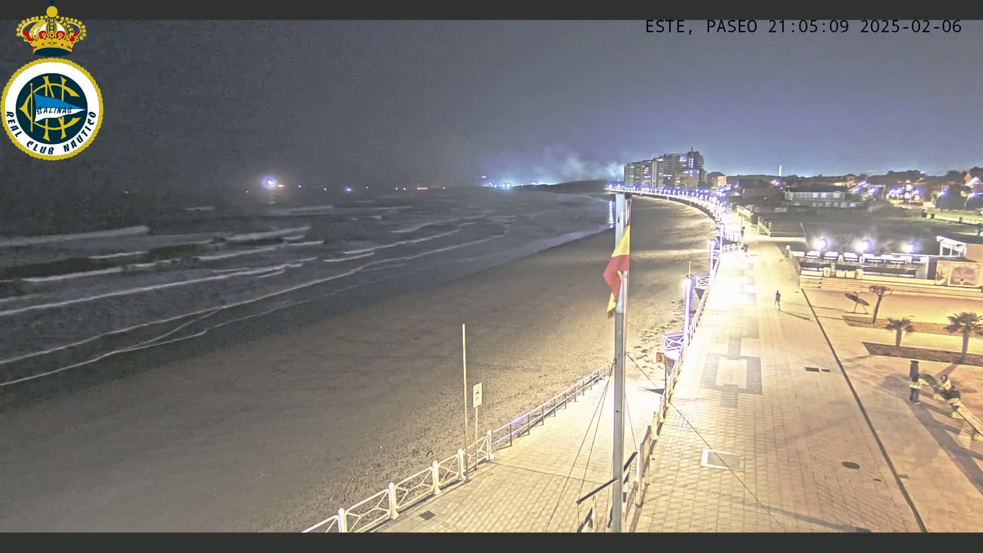 A nighttime view of a sandy beach with gently lapping waves, a paved boardwalk with lights, and buildings in the distance under a dark, clear sky.