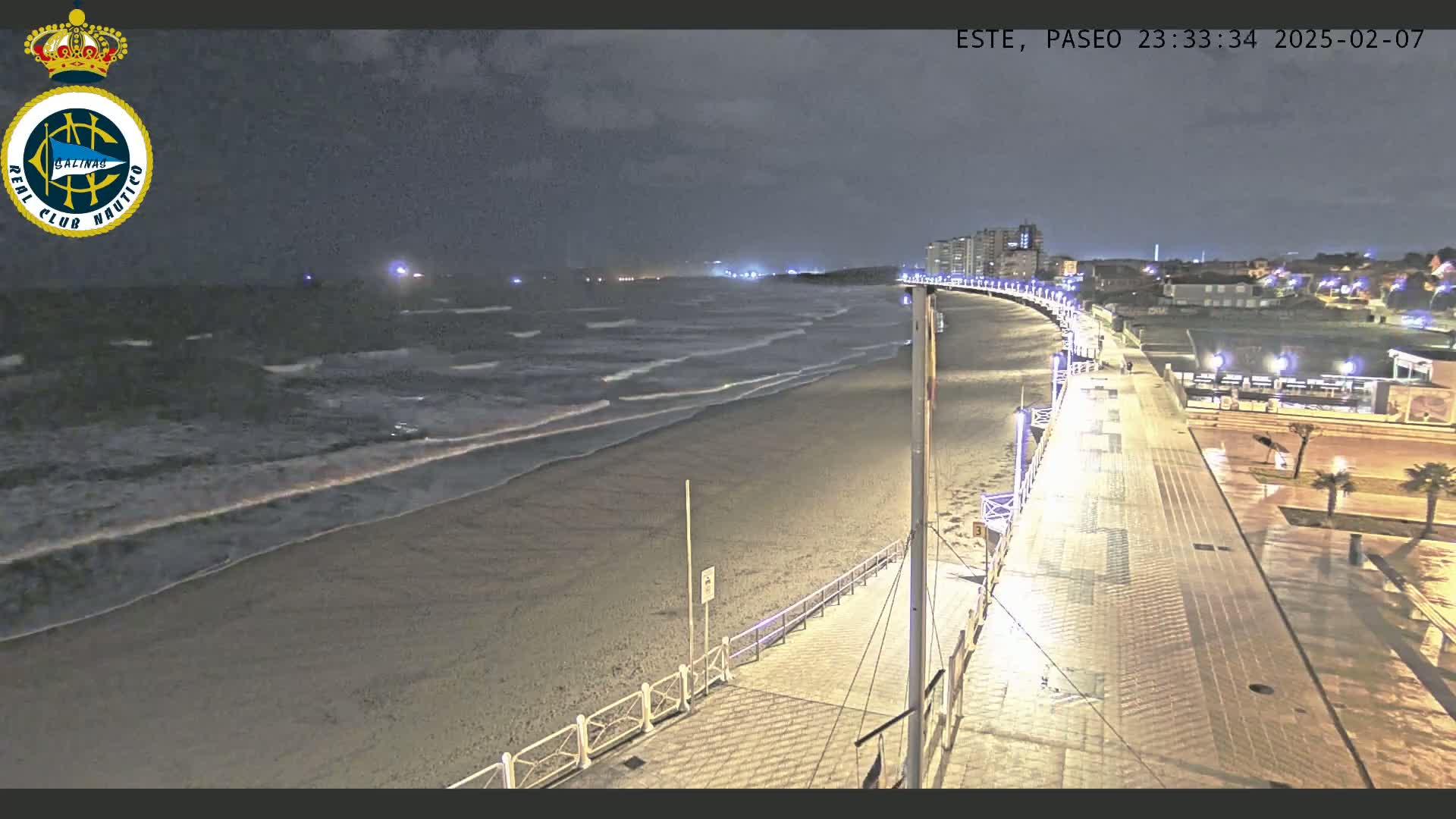 A nighttime beach scene shows moderate waves lapping a sandy shore, with a well-lit boardwalk curving alongside it toward buildings in the distance.