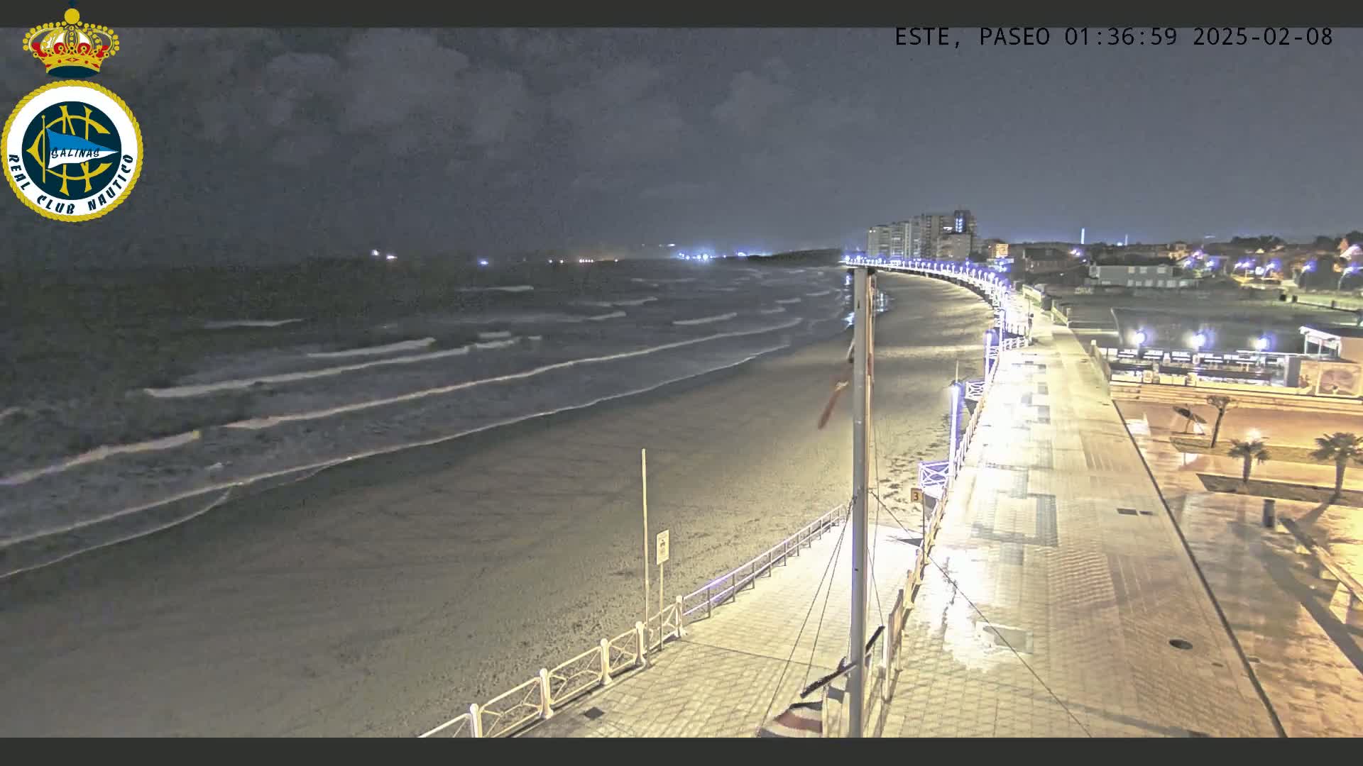 A nighttime view of a beach with gently lapping waves, a paved boardwalk with lights, and buildings in the distance under a partly cloudy sky.