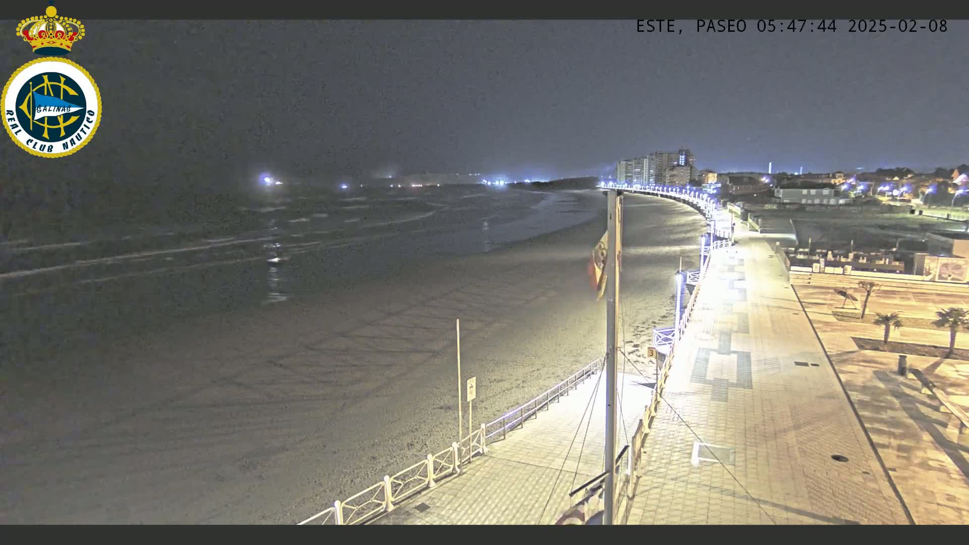 A nighttime view of a mostly empty beach with gently lapping waves, a paved walkway lined with lights, and buildings visible in the distance under a dark sky.