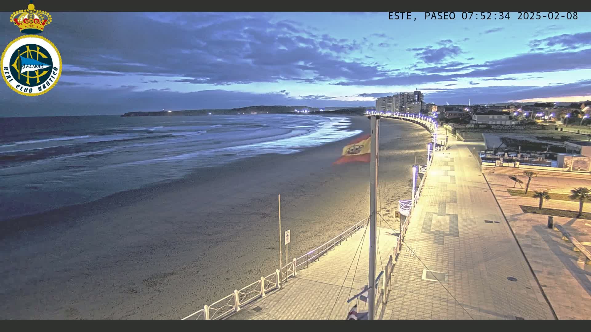 A beach at twilight, with a paved walkway lined with lights leading to buildings, shows a calm ocean with small waves under a cloudy sky.