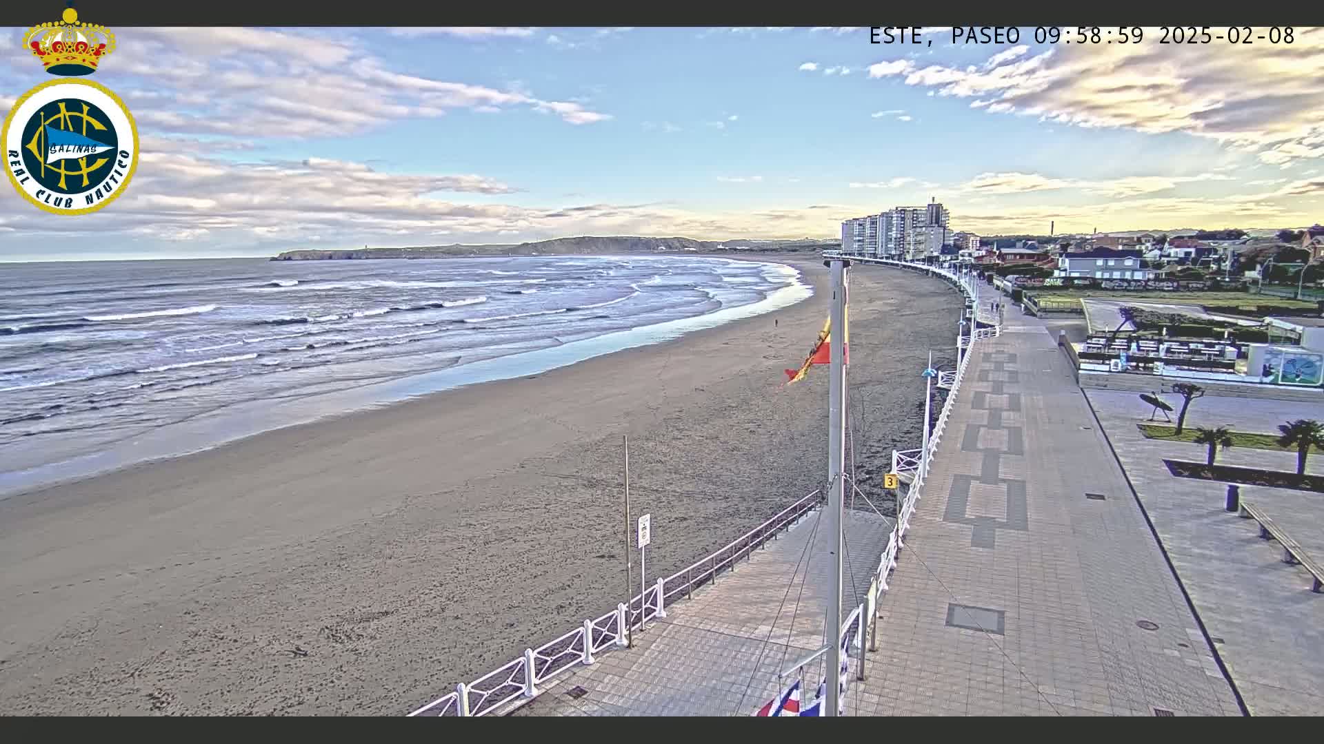 A mostly sunny day at a beach with moderate waves, showing a sandy beach, a paved boardwalk with buildings in the background.
