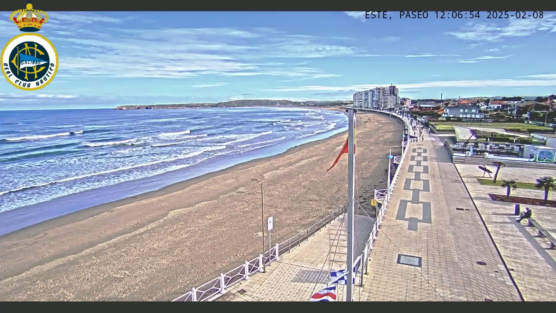 A sandy beach with moderate waves is next to a paved walkway and buildings under a mostly sunny sky.