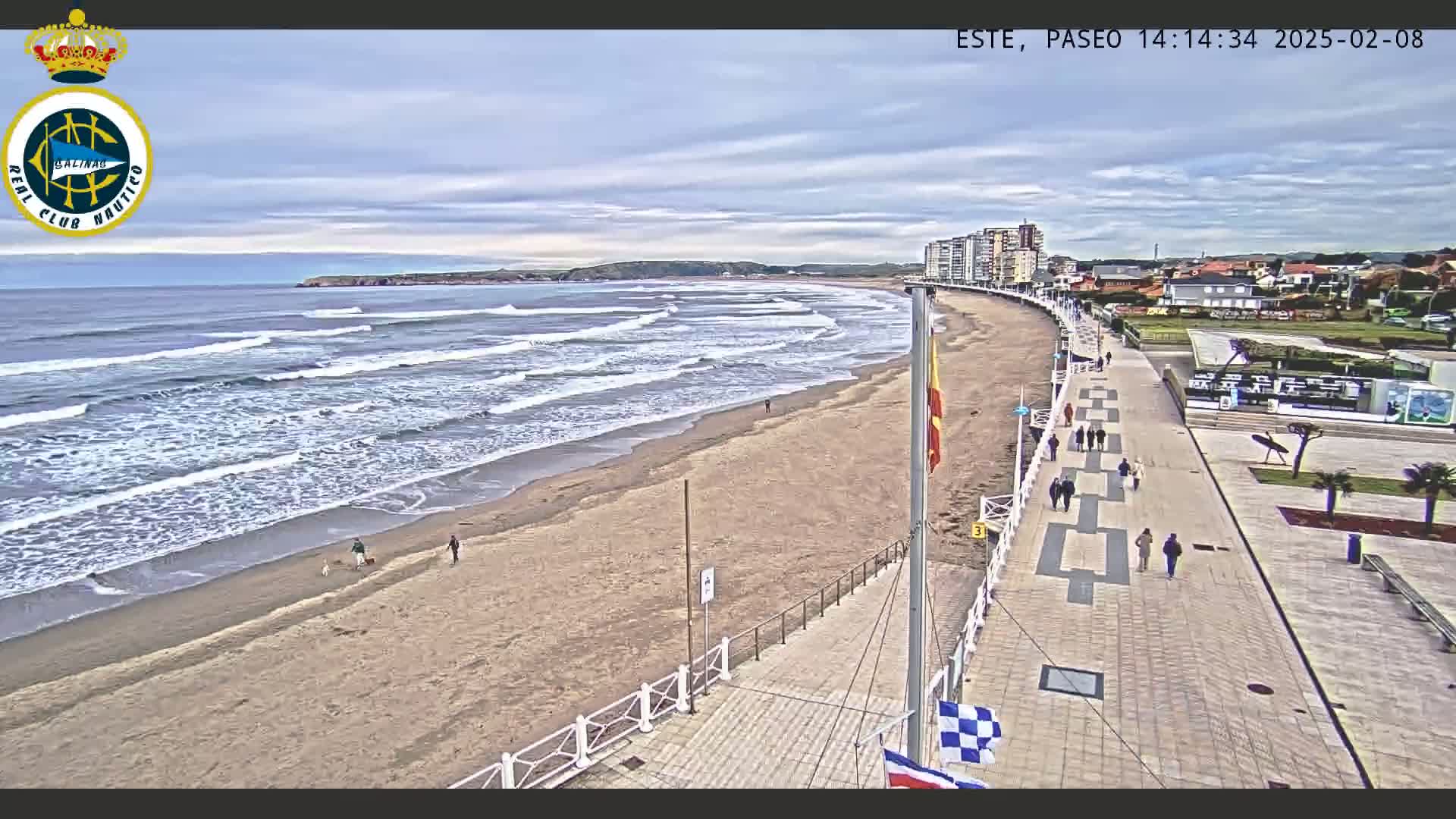 A sandy beach with gentle waves under a mostly cloudy sky is bordered by a paved walkway with pedestrians, and buildings are visible in the distance.