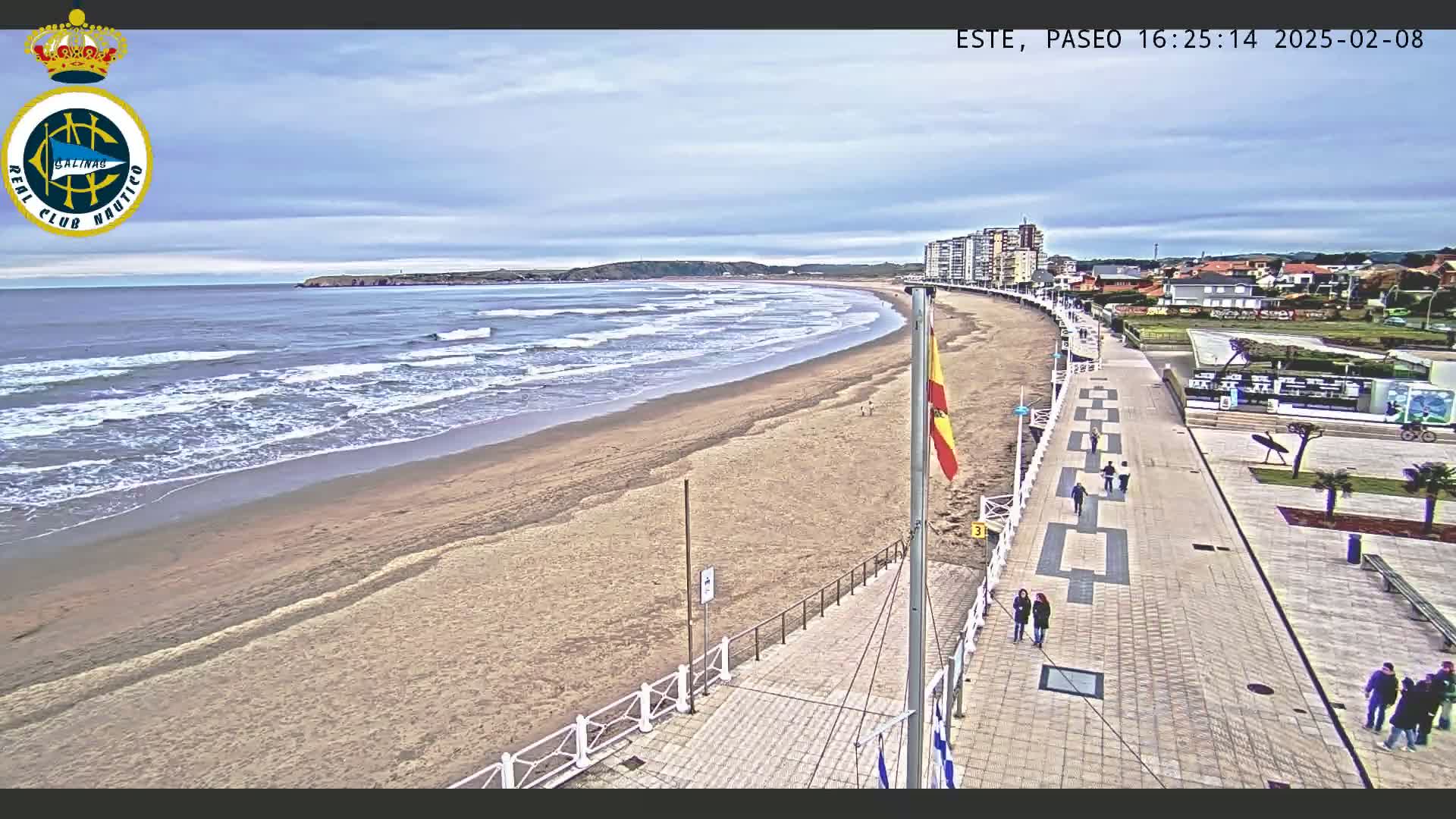 A sandy beach with gentle waves under a partly cloudy sky is bordered by a paved boardwalk with a few pedestrians, and buildings are visible in the background.