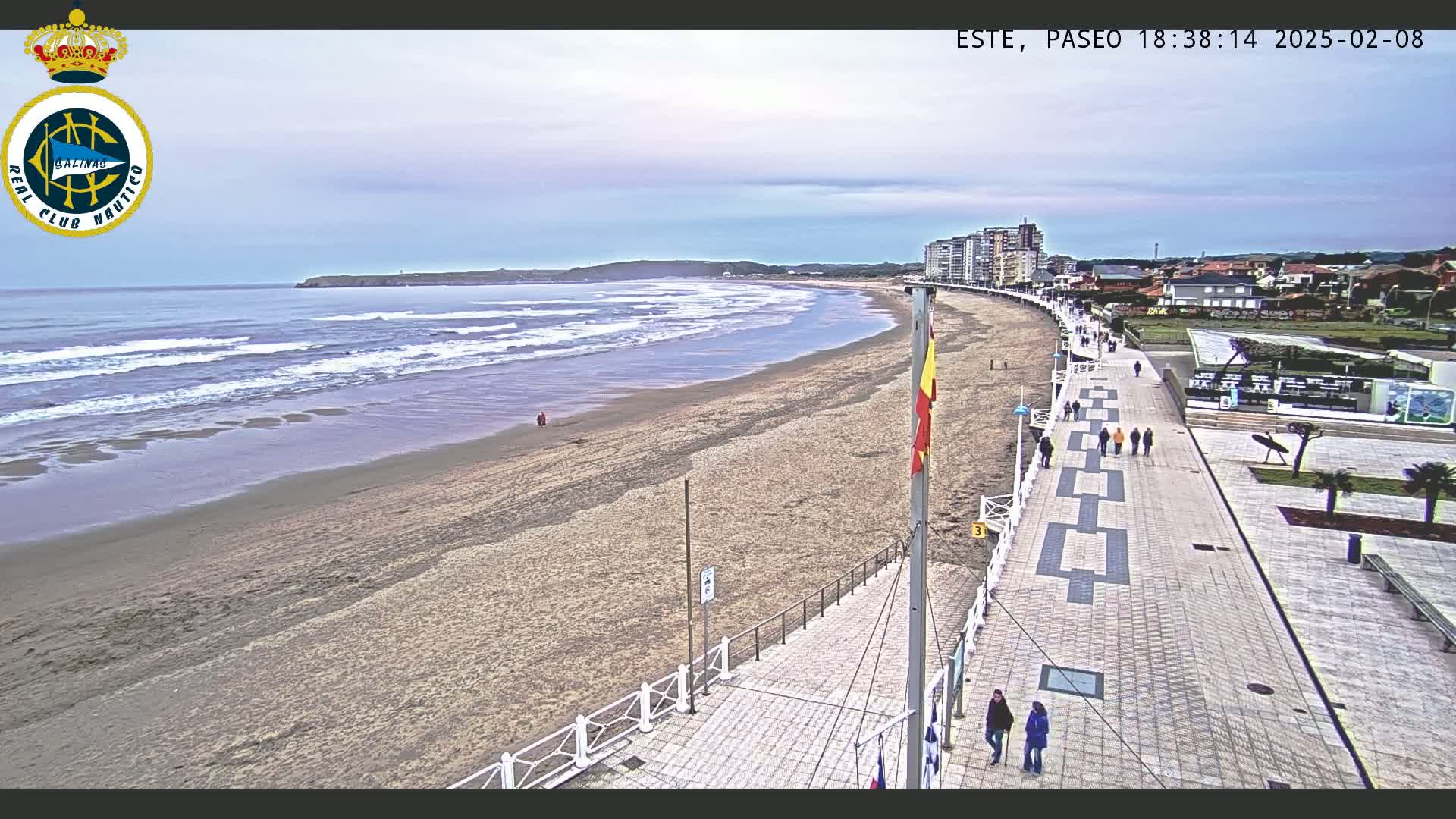 A sandy beach with gentle waves stretches alongside a paved walkway lined with people, under an overcast sky.