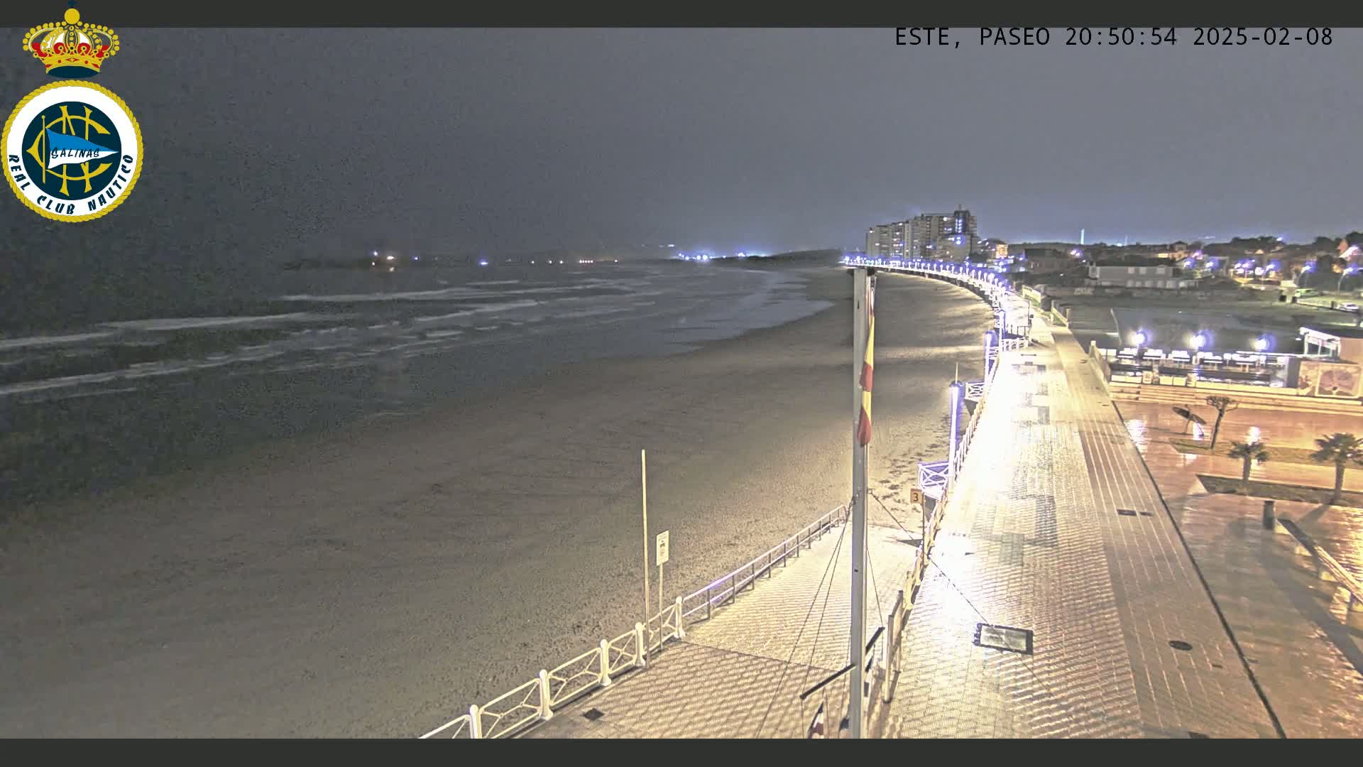A nighttime view of a mostly empty beach and boardwalk under a dark, overcast sky with a few lights visible along the shoreline and in the distance.