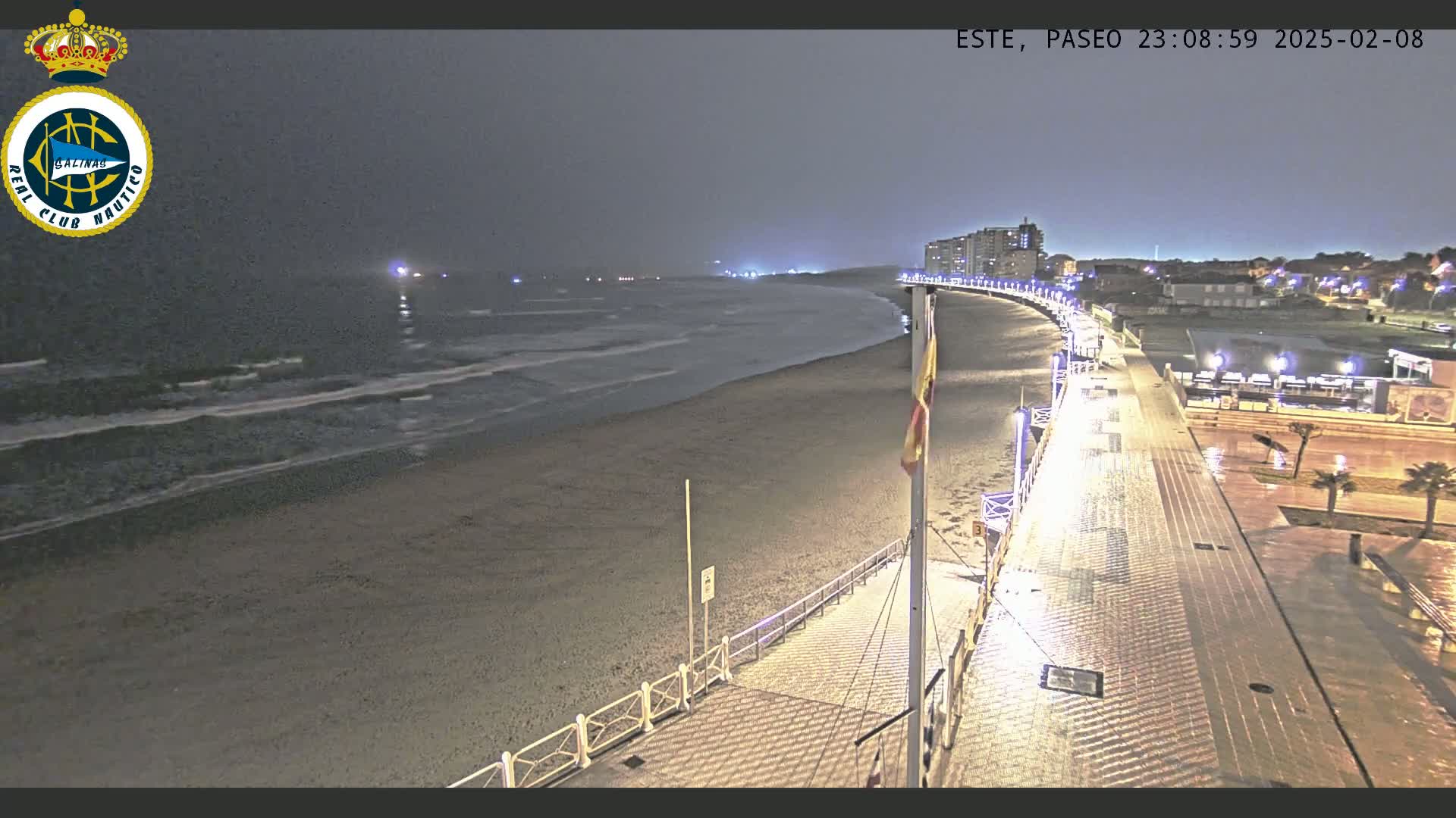 A nighttime view of a beach and boardwalk with gentle waves, under a dark, overcast sky.