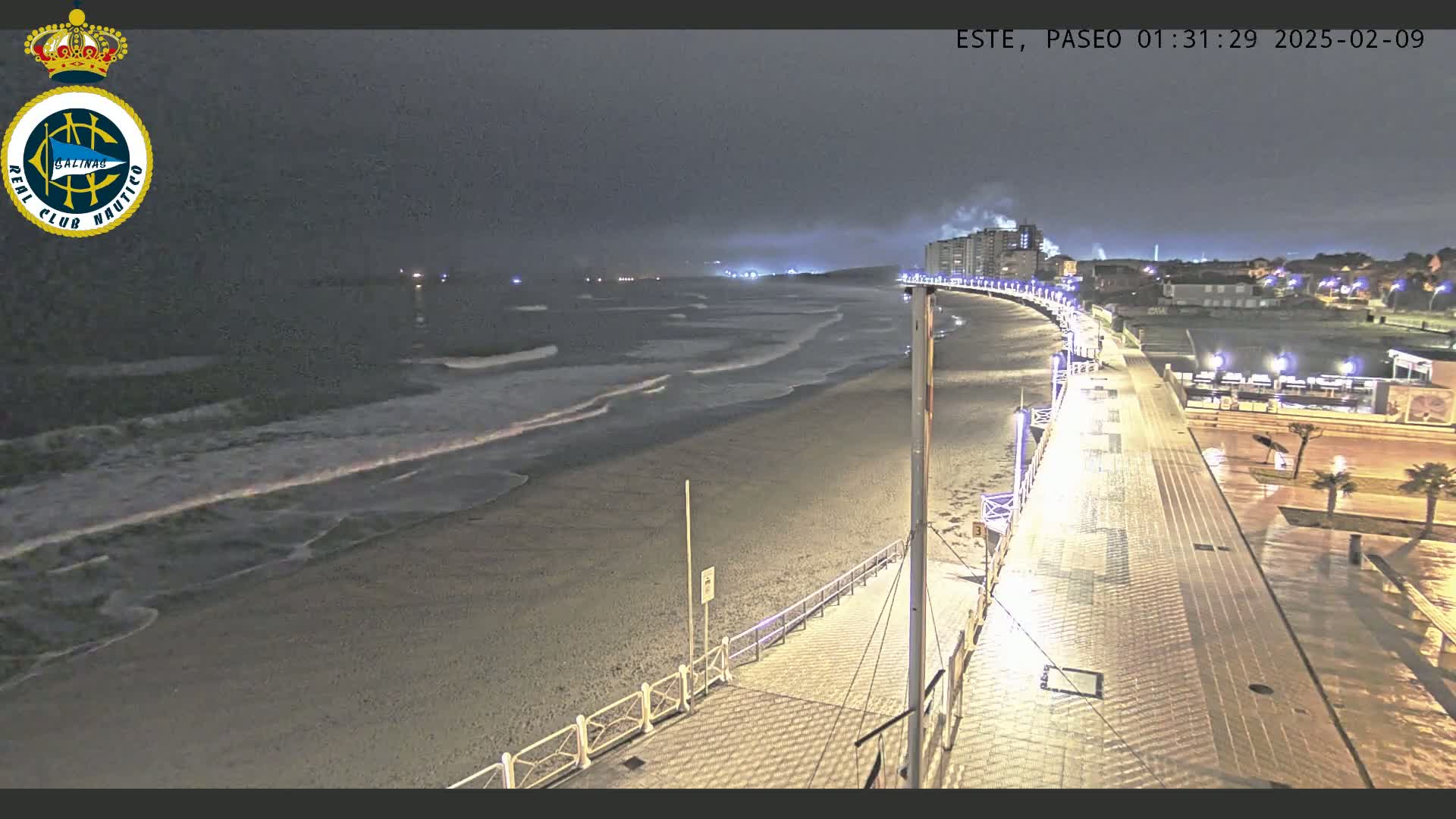 A nighttime view of a beach with gentle waves under a dark, overcast sky, showing a paved walkway curving along the shoreline, lined with lights, and buildings visible in the distance.