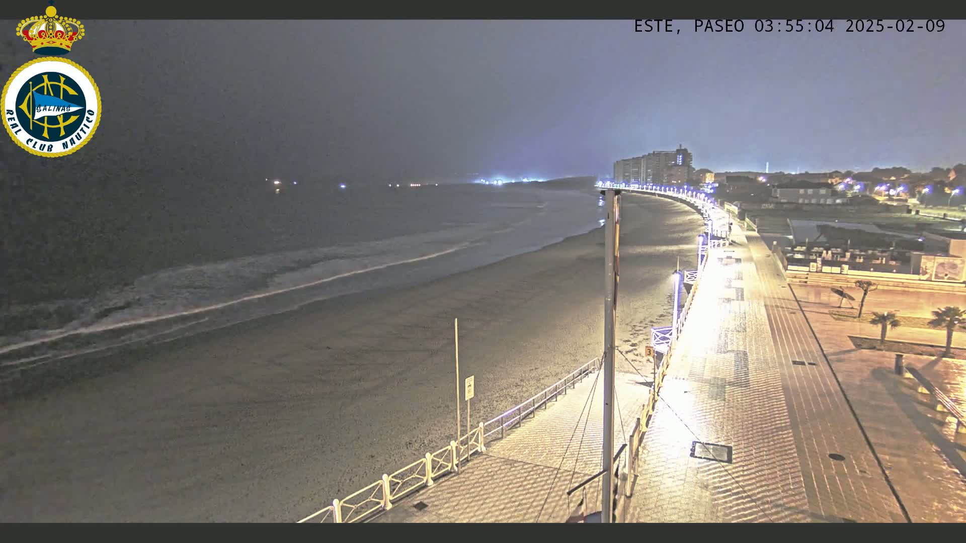 A nighttime, dimly lit beach scene shows a paved walkway curving along the shoreline next to a dark sandy beach with gentle waves under an overcast sky.