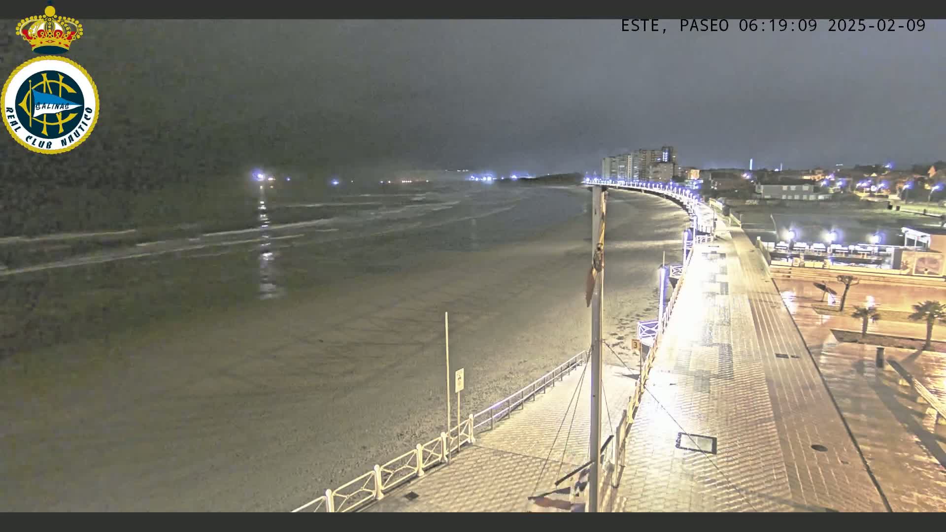 A nighttime view of a wet, dark beach and boardwalk with calm waves and distant lights.