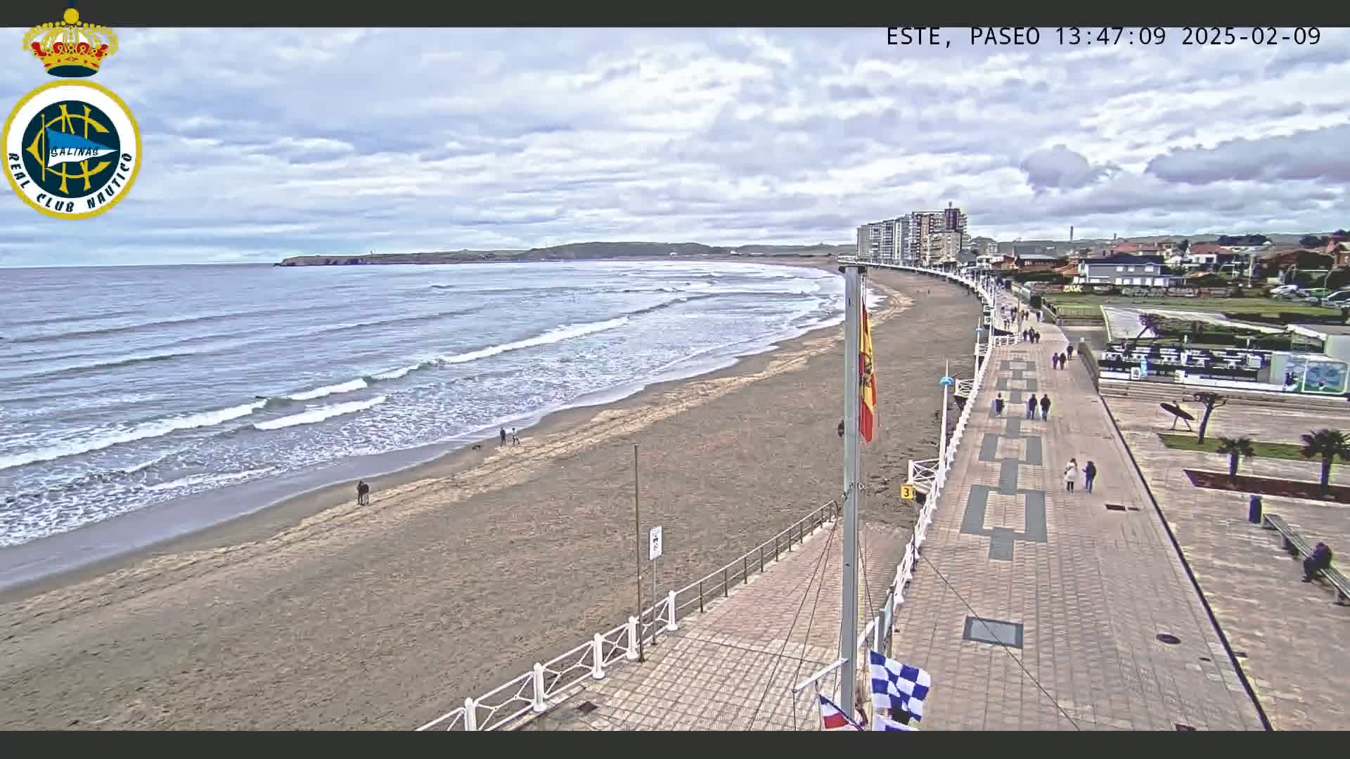 A sandy beach with gentle waves under a partly cloudy sky is bordered by a paved promenade with pedestrians, and buildings are visible in the distance.