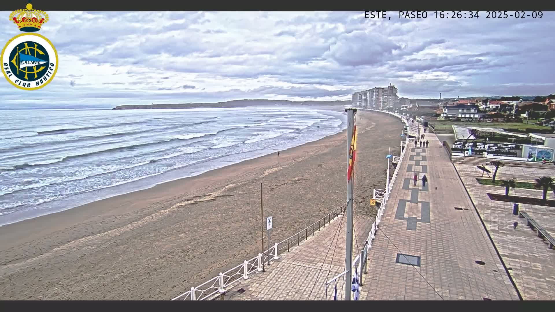 A mostly cloudy day at a beach with a wide sandy shore, moderate waves, and a paved boardwalk with people walking along it, next to buildings.