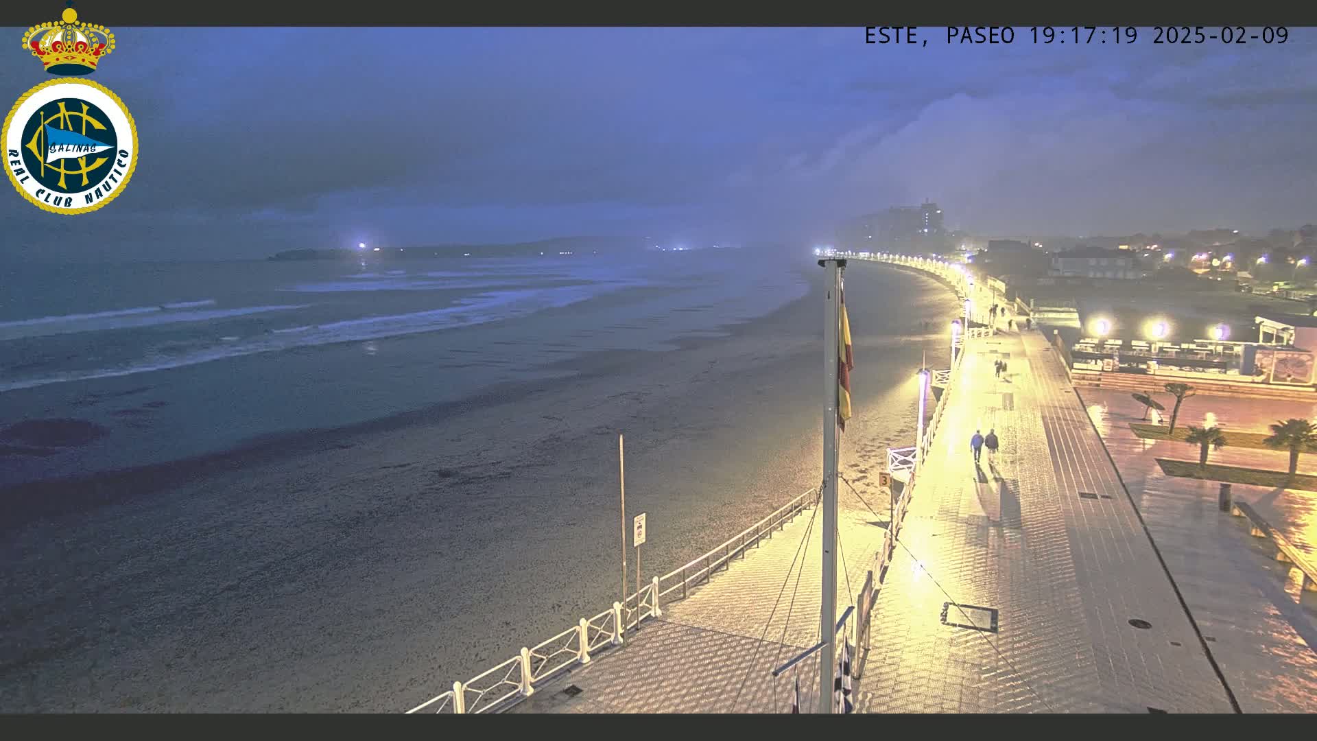 A nighttime beach scene shows a wet boardwalk overlooking a dark, sandy beach and a calm ocean under a cloudy sky.