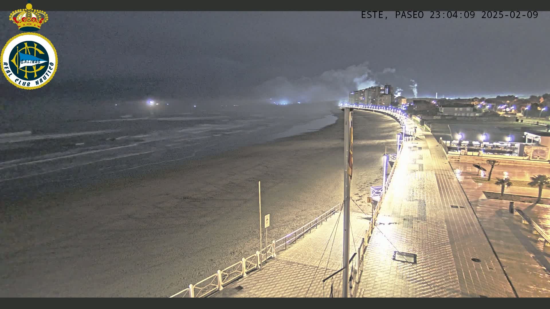 A nighttime view of a mostly empty beach and boardwalk, with mild waves and a hazy atmosphere.