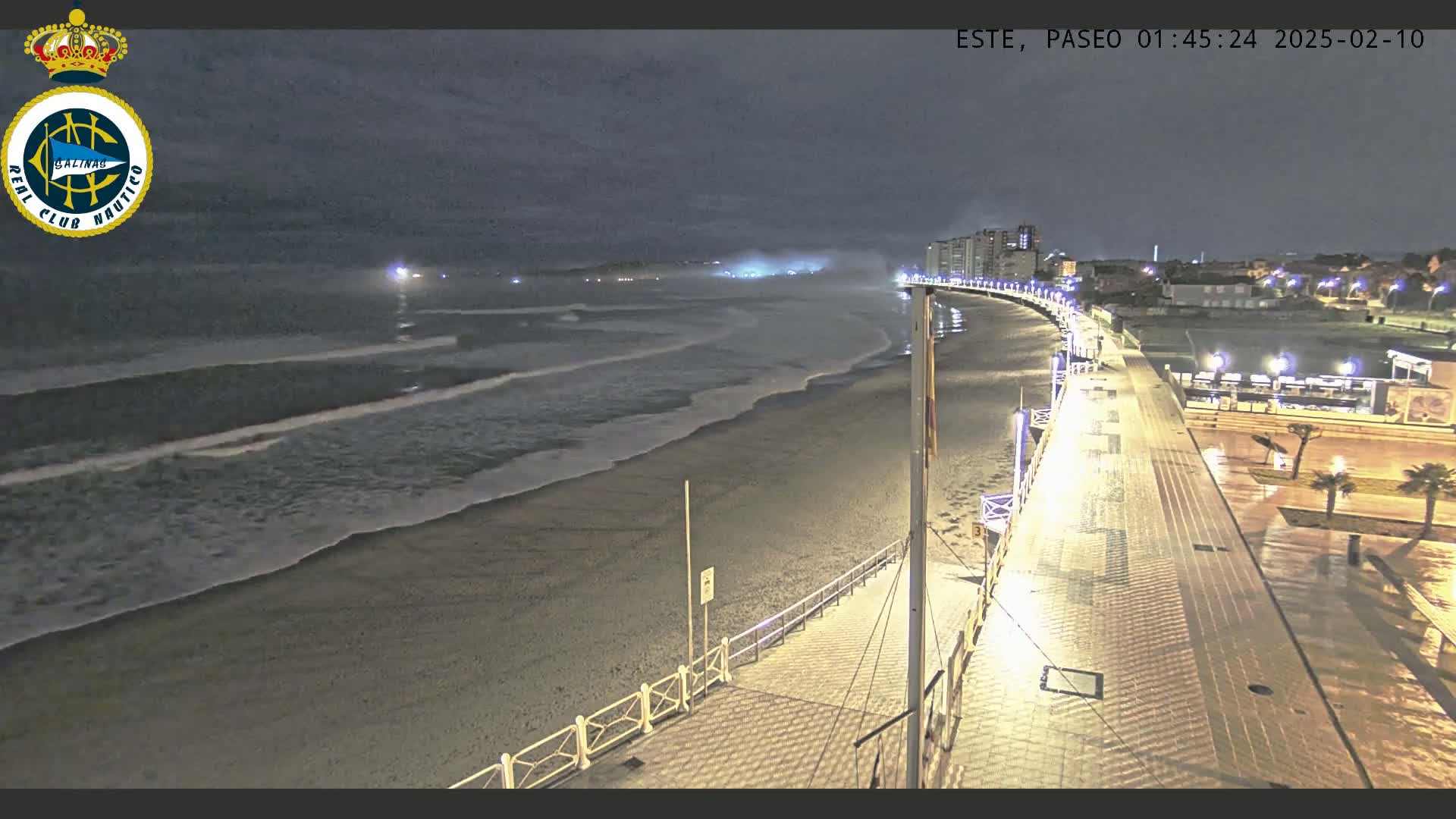 A nighttime view shows a dimly lit beach with gentle waves rolling onto the shore, next to a paved boardwalk with lights, under a hazy sky.