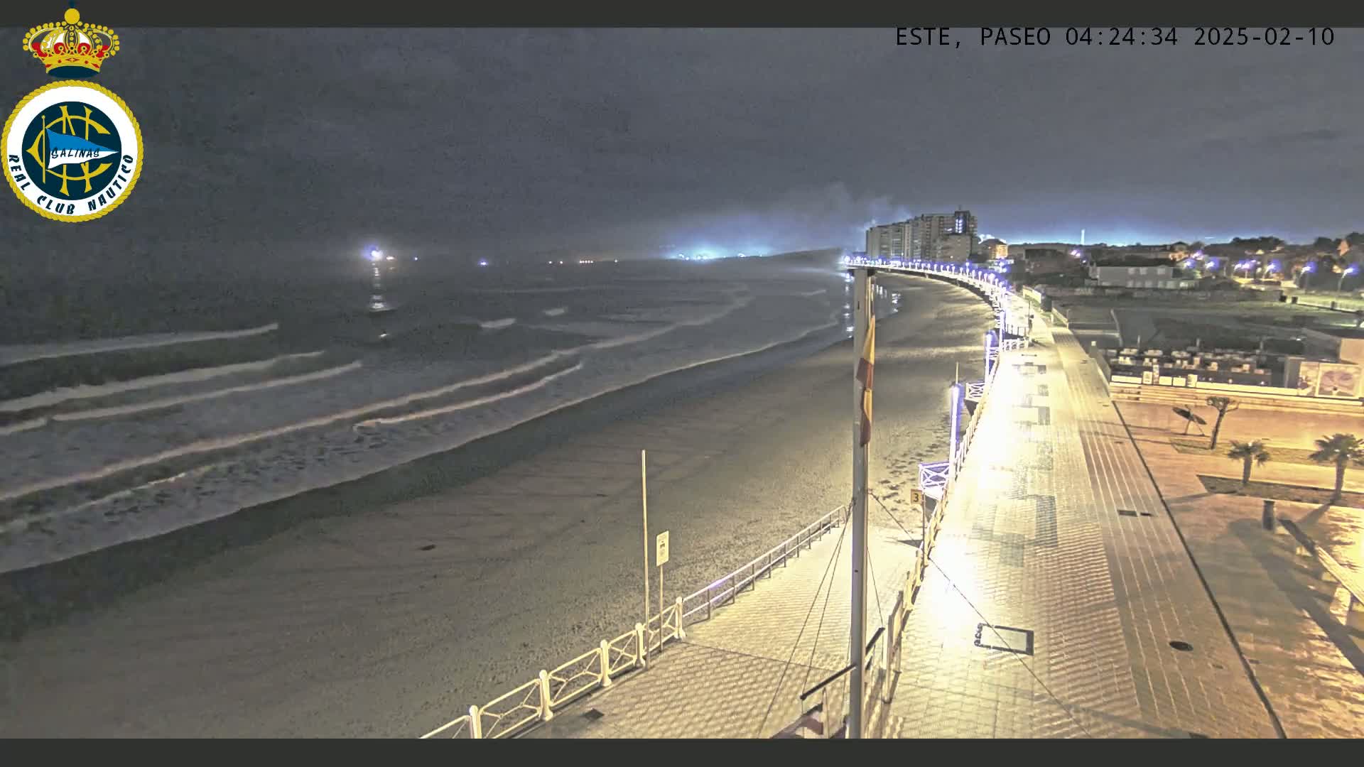 A nighttime view of a mostly empty beach and boardwalk with gentle waves under a dark sky.