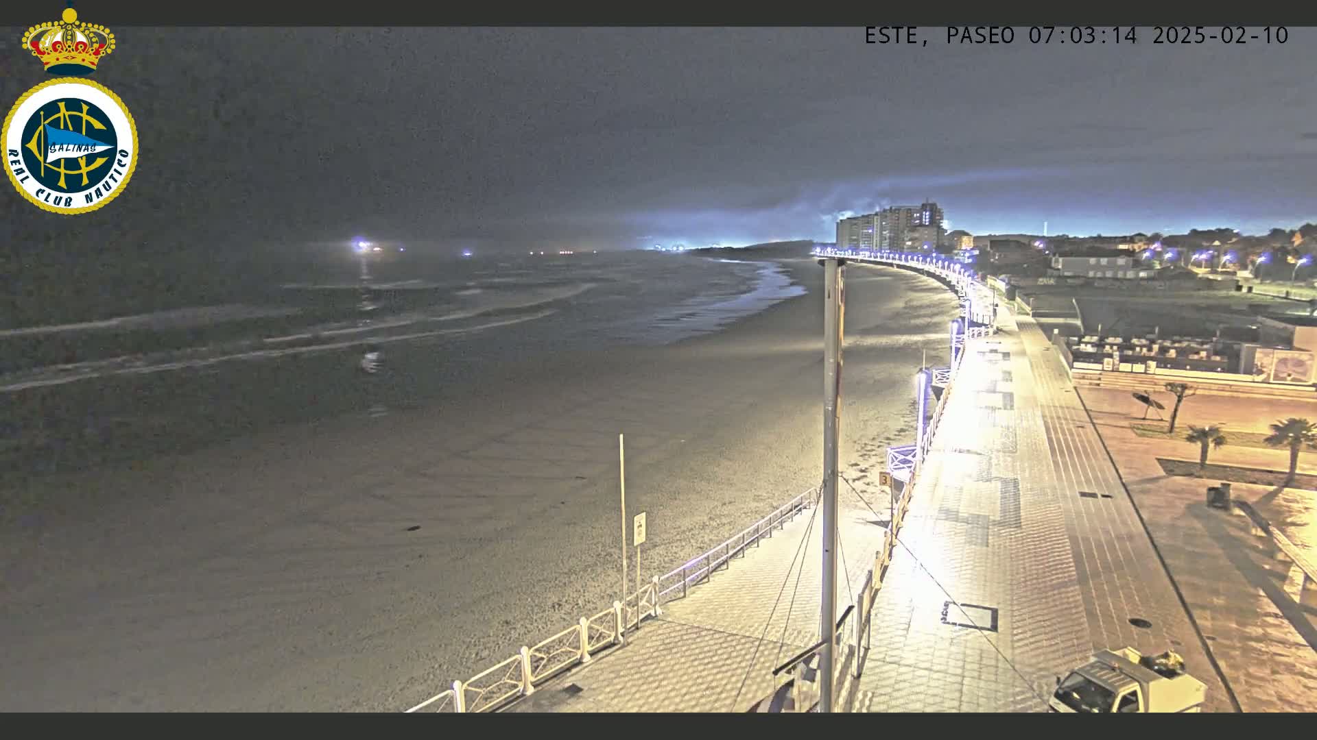 A nighttime beach scene shows a paved walkway curving along a sandy beach under a dark, cloudy sky, with lights illuminating the walkway and distant buildings.