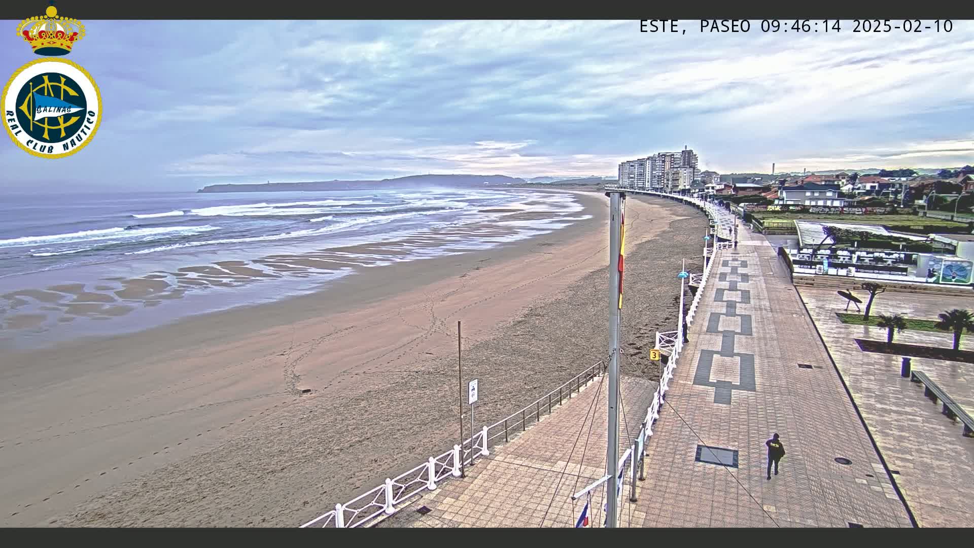 A sandy beach with moderate waves under a cloudy sky is next to a paved walkway lined with buildings and a few people walking.
