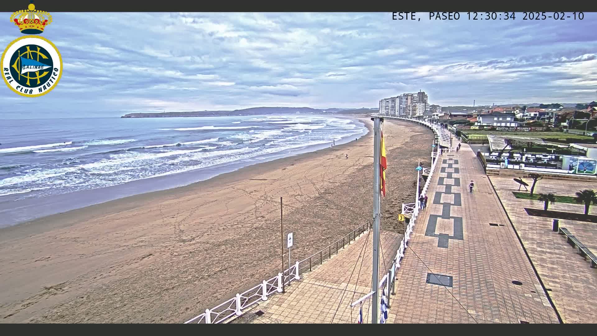 A sandy beach with gentle waves under a cloudy sky is adjacent to a paved boardwalk lined with buildings, leading to a plaza with landscaping.