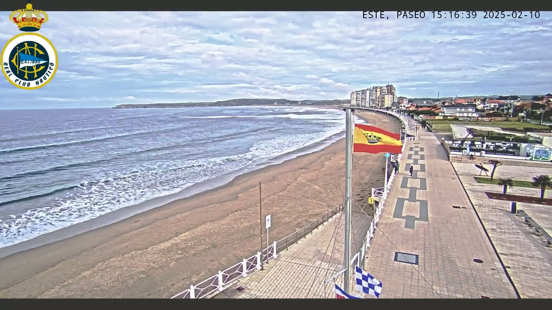 A sandy beach with gentle waves under a partly cloudy sky is bordered by a paved walkway and buildings.