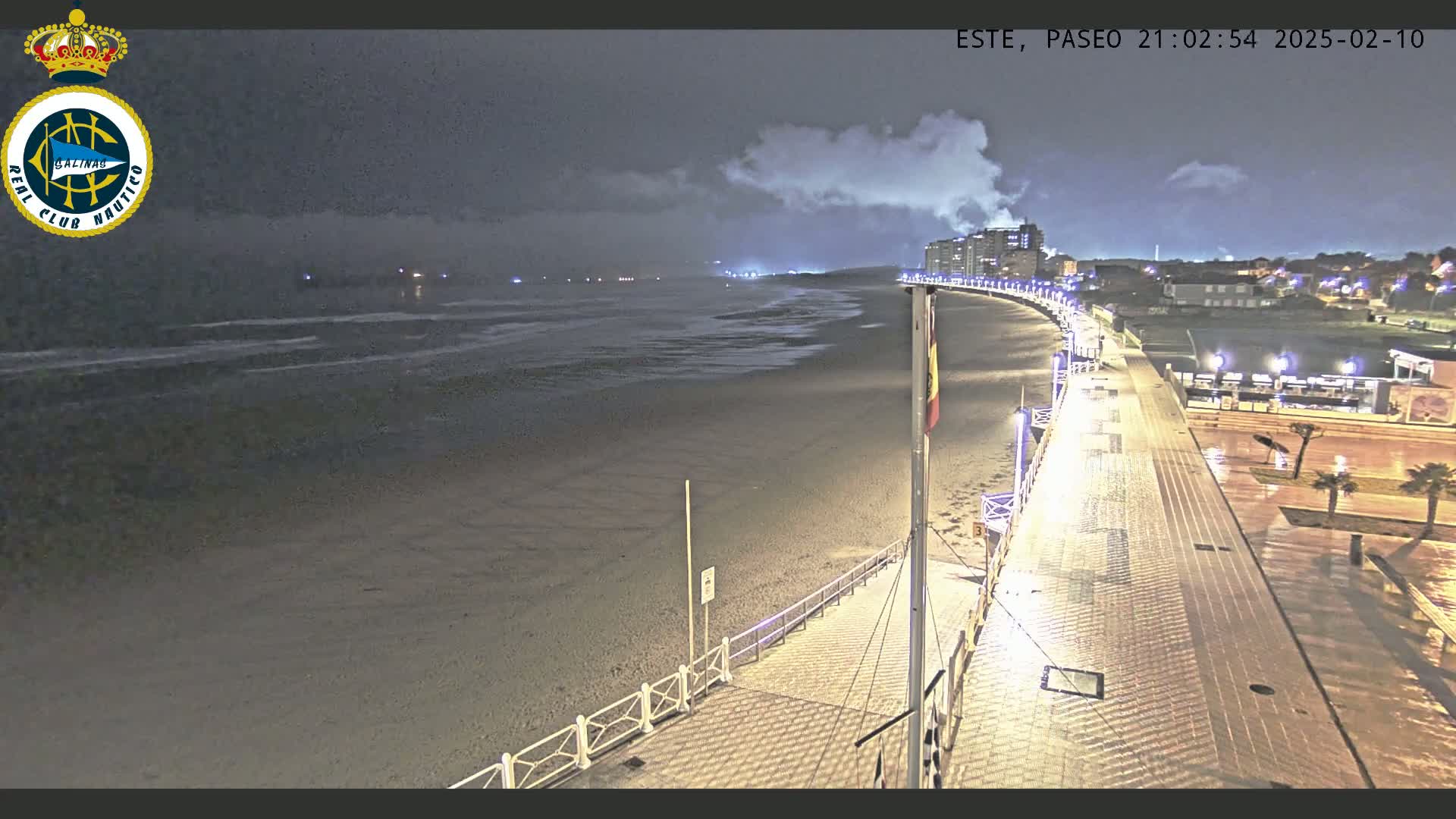 A nighttime view of a mostly empty beach and boardwalk, under a cloudy sky with light rain.