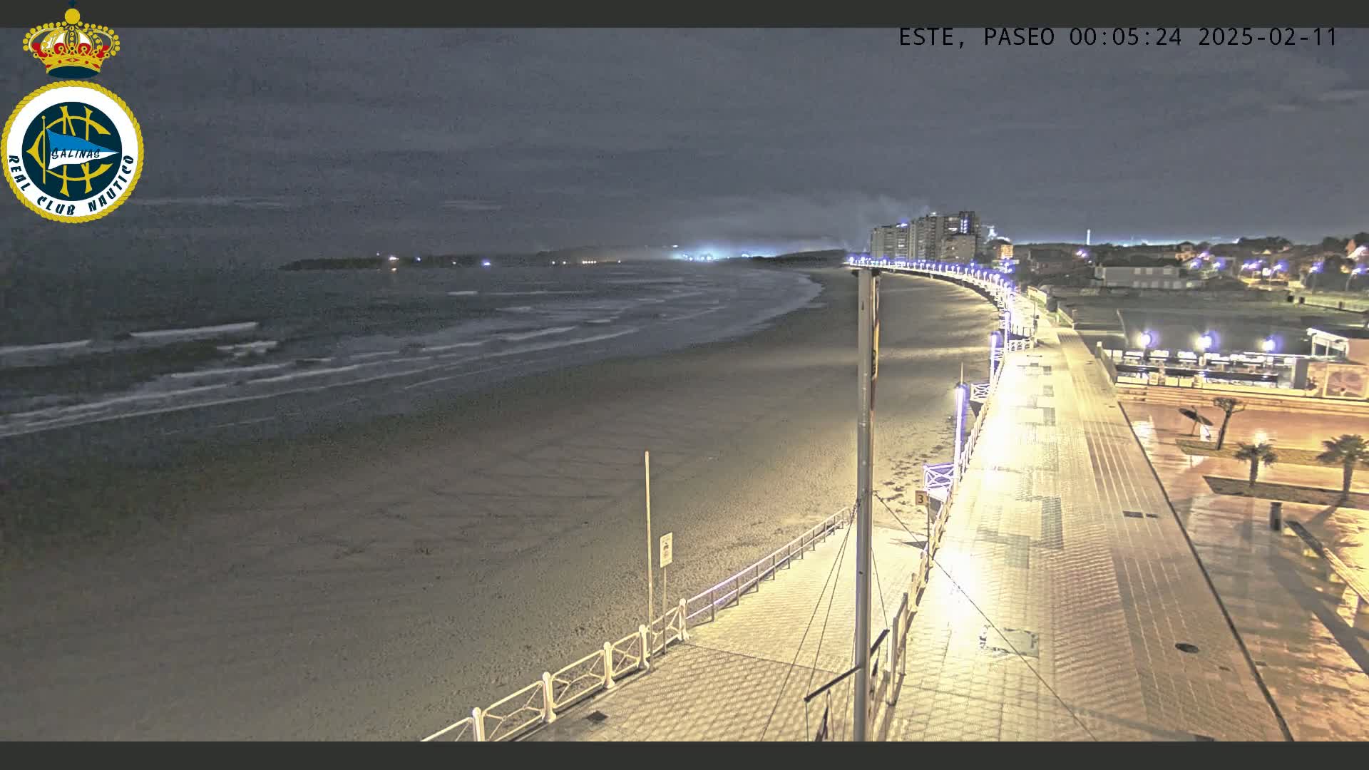 A nighttime view of a mostly empty beach and illuminated boardwalk, with gentle waves lapping the shore under a dark, cloudy sky.