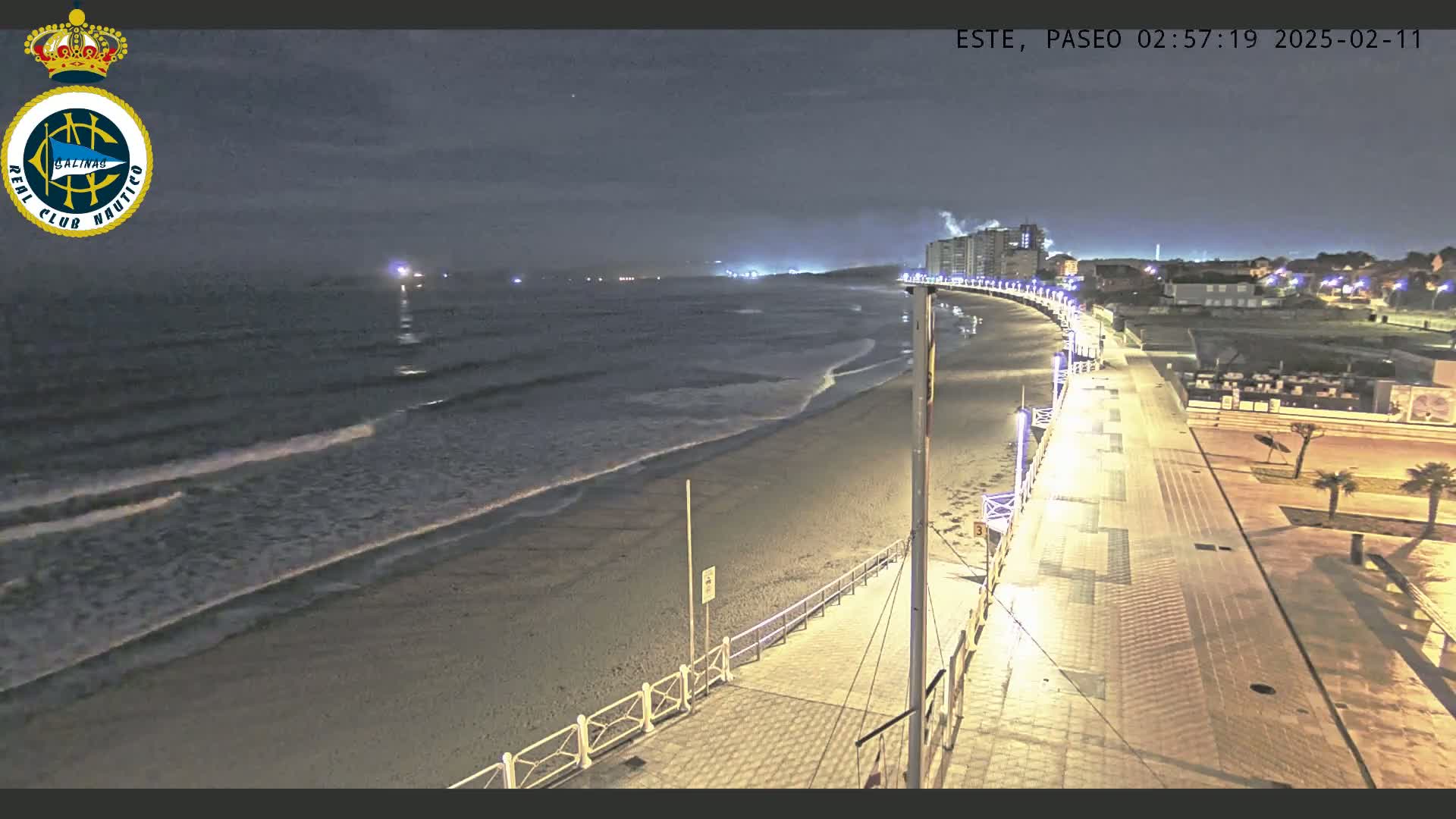 A nighttime view of a sandy beach with gentle waves lapping the shore, a paved walkway lined with lights, and buildings in the distance under a dark sky.