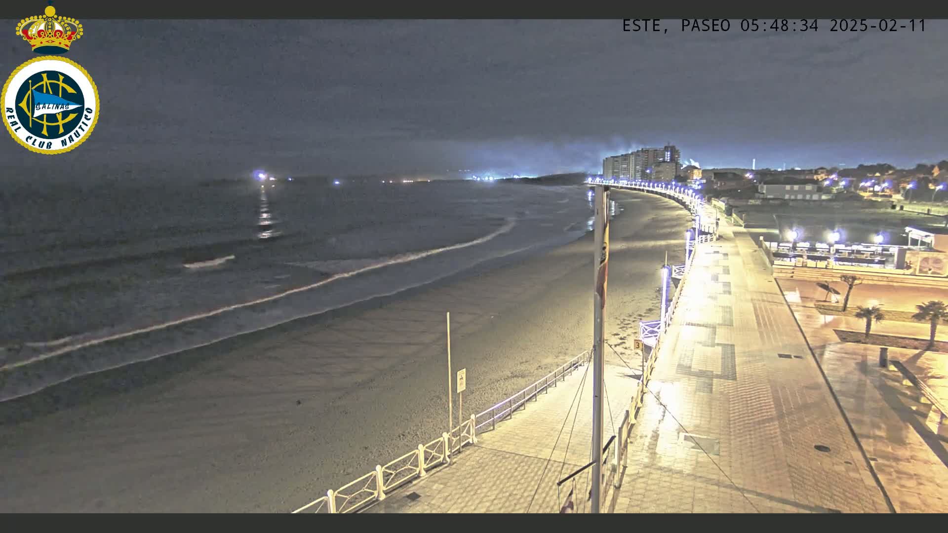 A nighttime view of a beach with gentle waves lapping the shore, a paved boardwalk lined with lights, and buildings in the distance under a partly cloudy sky.