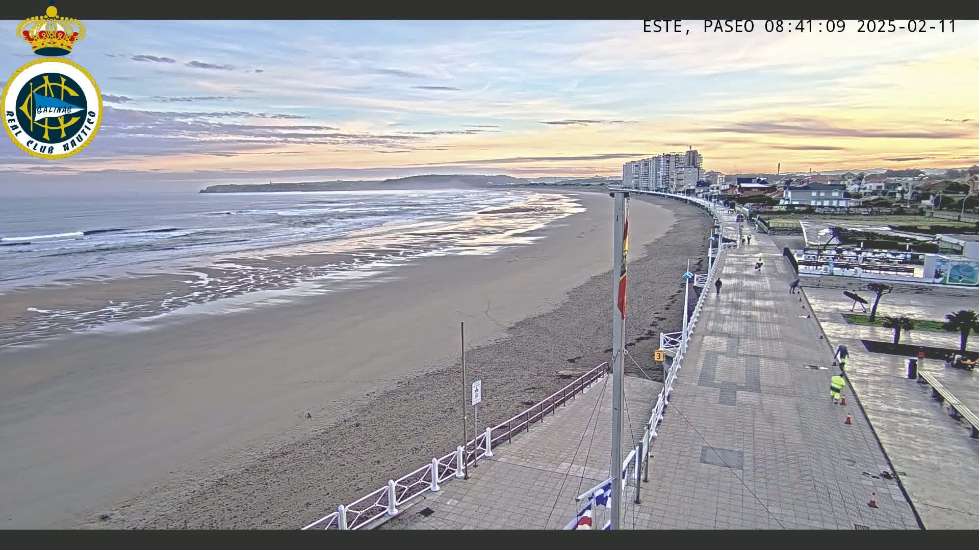 A sandy beach with gentle waves under a partly cloudy sunset sky is seen from above, alongside a paved promenade with people walking and workers cleaning.