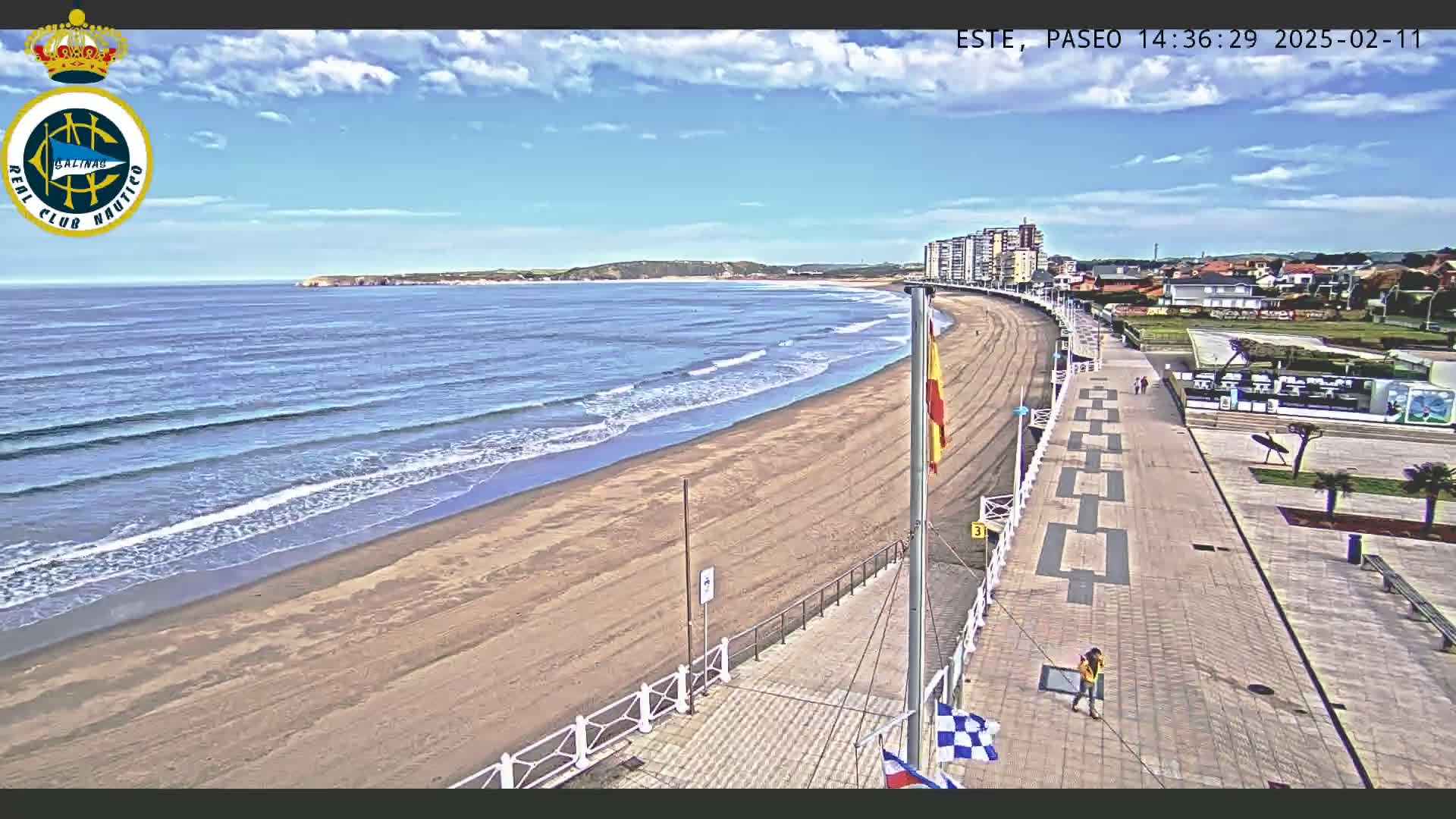 A sandy beach with gentle waves under a mostly sunny sky is bordered by a paved walkway and buildings.