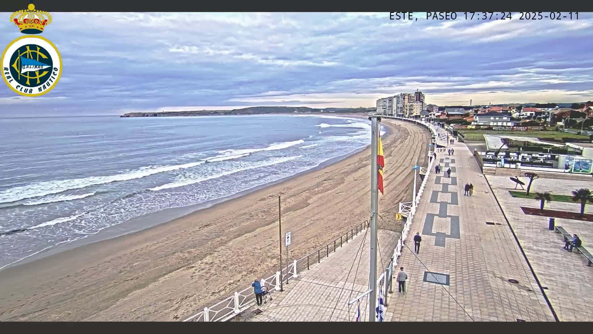 A sandy beach with gentle waves under a partly cloudy sky is bordered by a paved walkway with people strolling along it, leading to buildings in the distance.