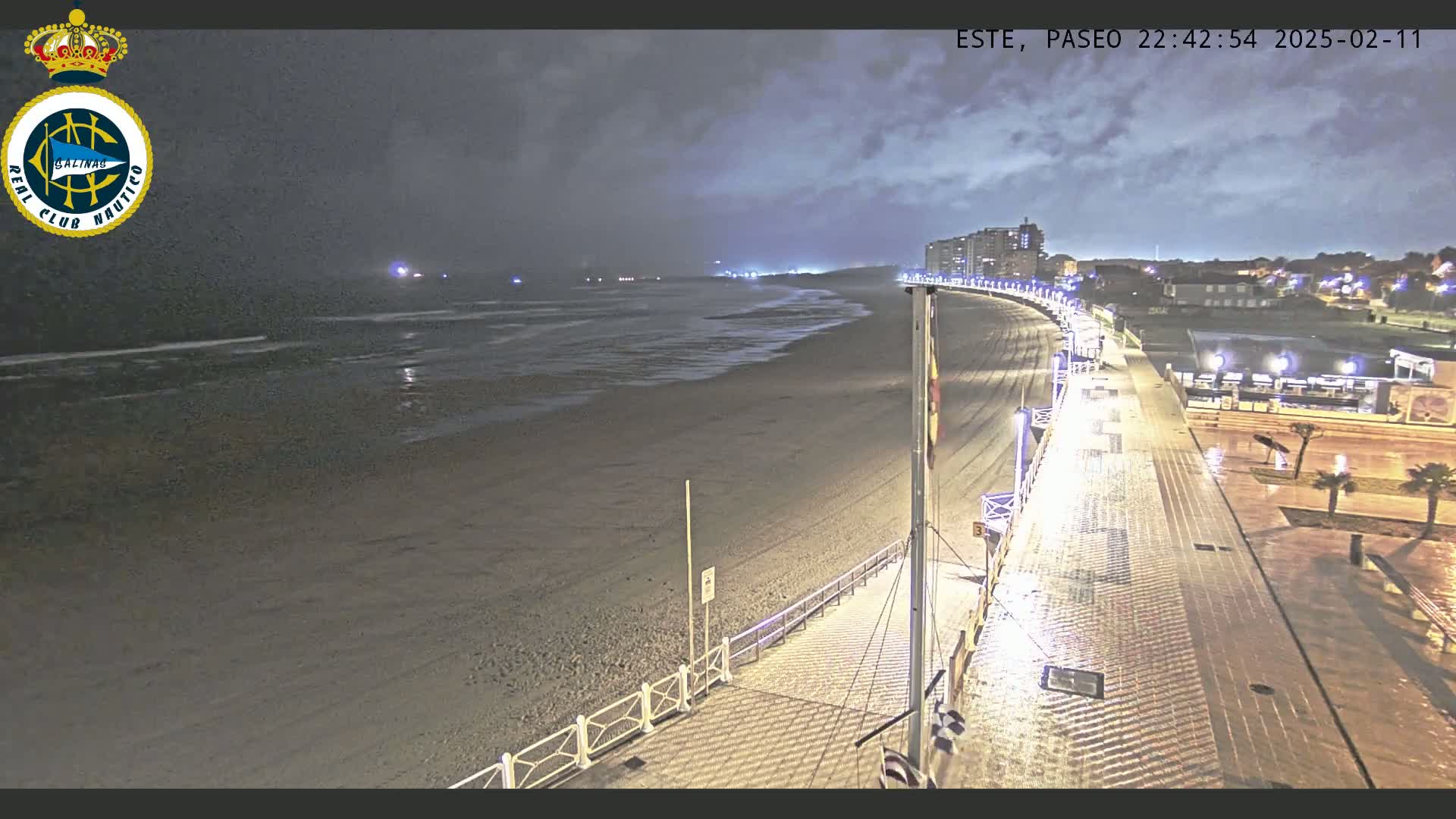 A nighttime view of a wet, sandy beach and a paved boardwalk, lined with lights and buildings in the background under a cloudy sky.