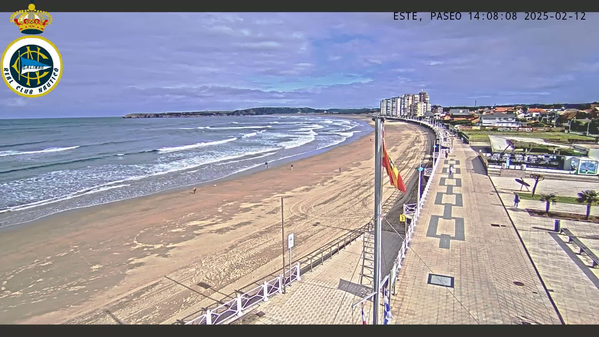 A sandy beach with gently rolling waves under a partly cloudy sky is bordered by a paved walkway and buildings.