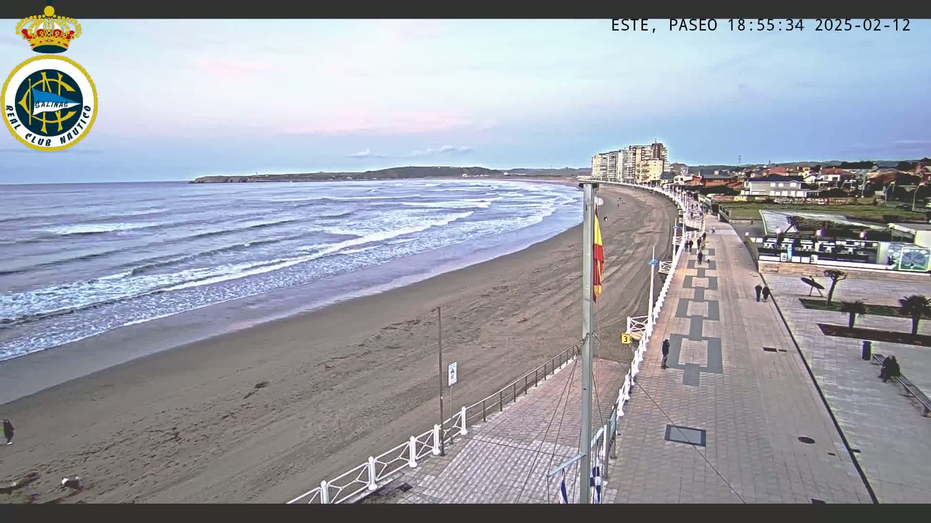 A sandy beach with moderate waves under a twilight sky is adjacent to a paved boardwalk with several people walking along it, and buildings are visible in the distance.