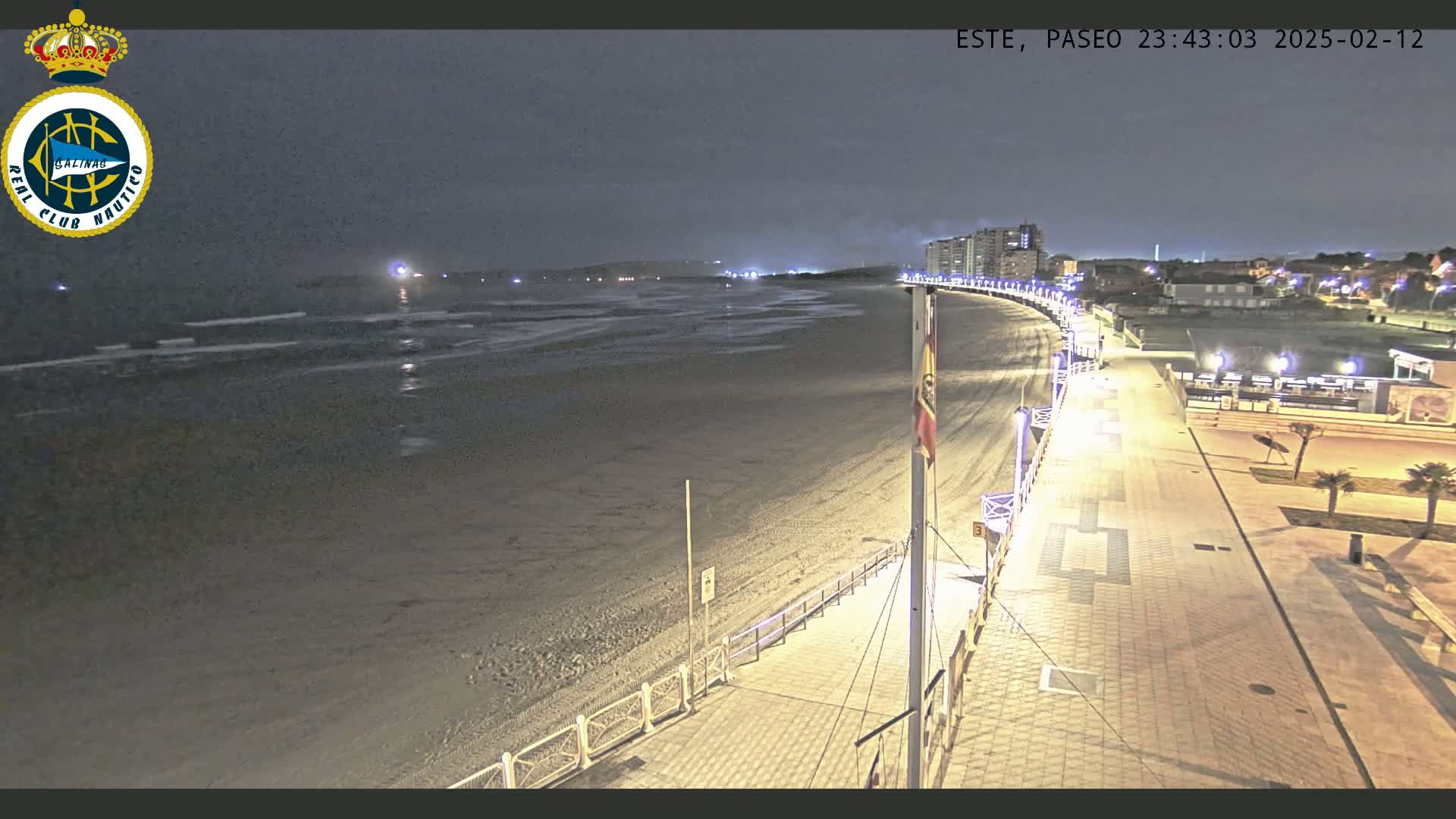 A nighttime view of a mostly empty beach and boardwalk, with gentle waves lapping the shore under a dark sky.