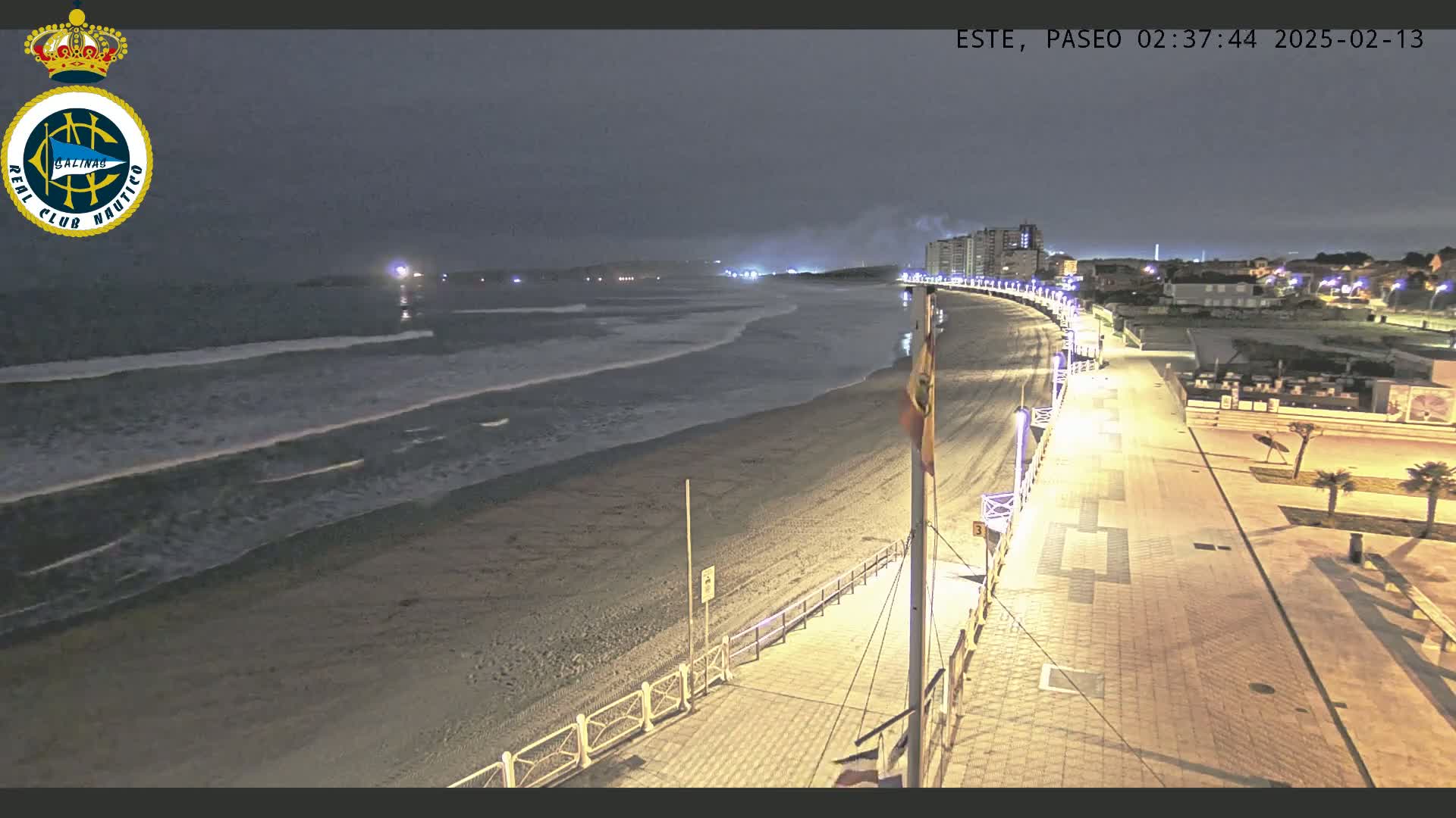 A nighttime view of a beach with gentle waves, a paved boardwalk, and buildings in the distance under a dark sky.