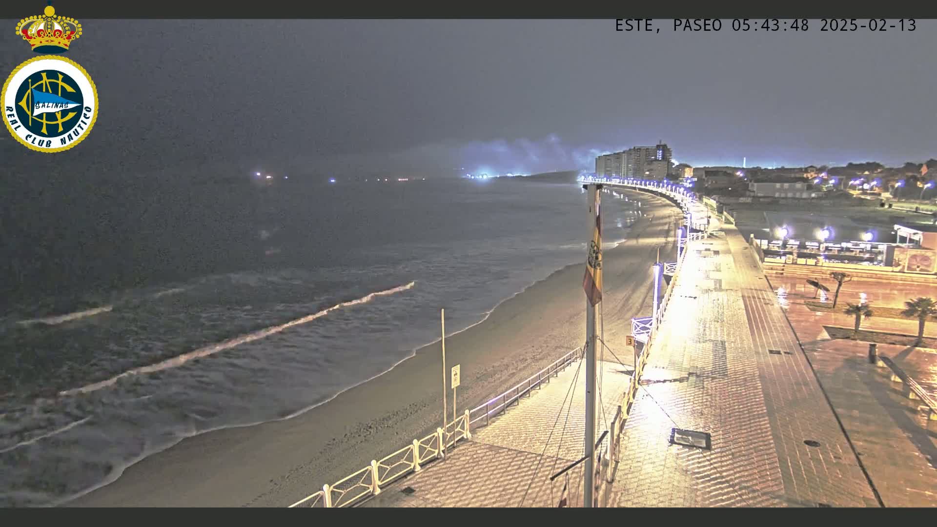 A nighttime view of a beach with gentle waves under a cloudy sky, showing a brightly lit boardwalk and buildings in the distance.