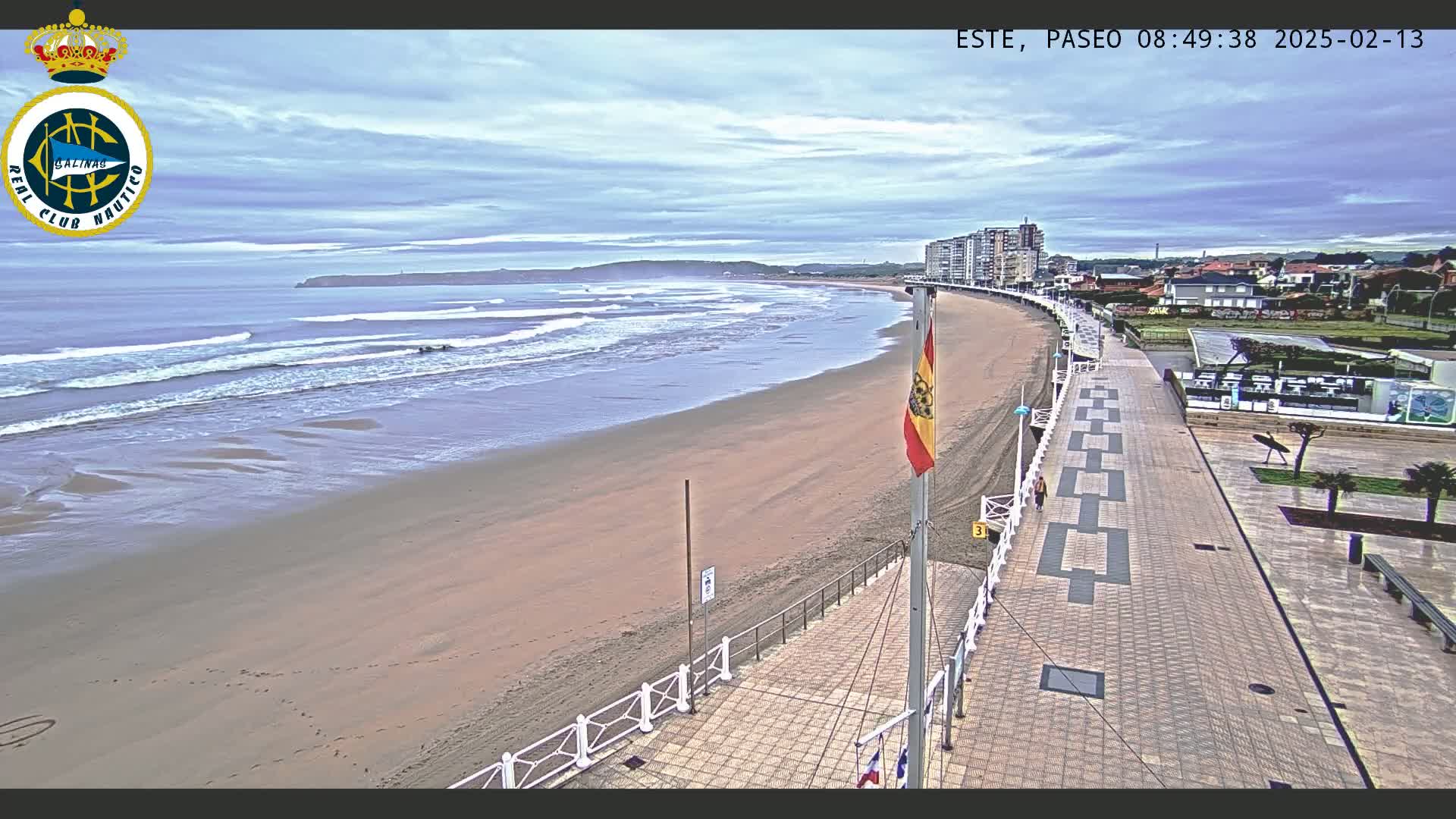 A sandy beach with gentle waves under a cloudy sky is bordered by a paved walkway leading to buildings, and a Spanish flag stands on the promenade.
