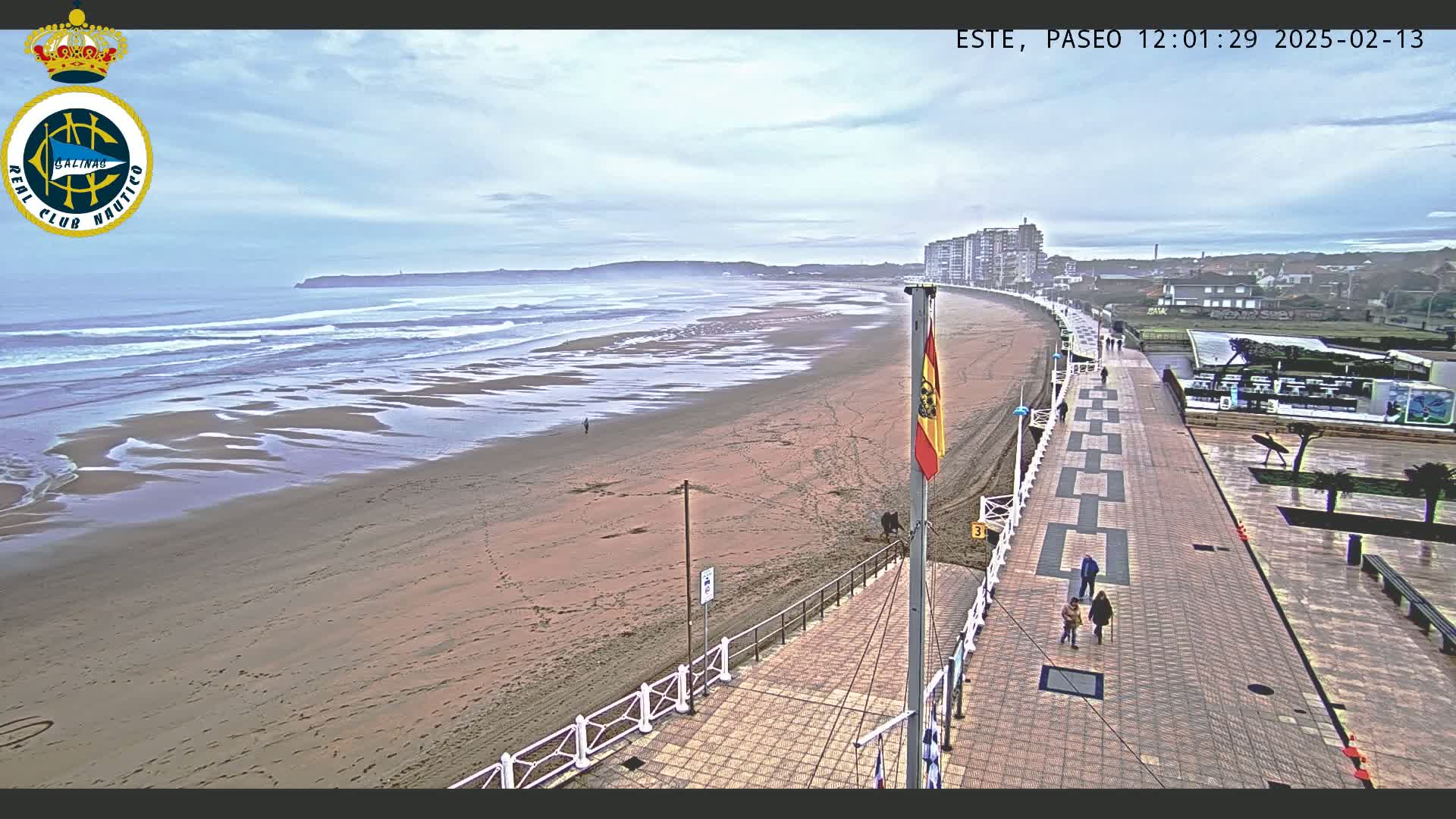 A sandy beach with gentle waves under an overcast sky is seen from above, alongside a paved boardwalk with people walking and buildings in the distance.