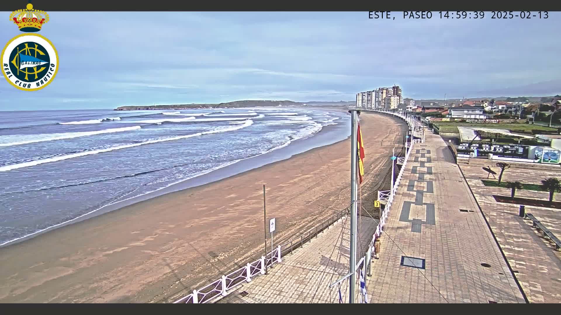 A sandy beach with waves rolling in under a partly cloudy sky is viewed from a paved boardwalk next to a row of buildings.