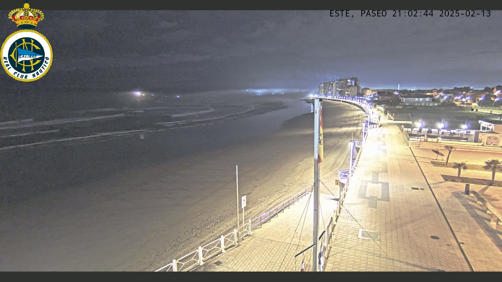 A nighttime view of a sandy beach with gentle waves under a mostly clear sky, showing a paved boardwalk with lights extending from a beachfront building toward the water.