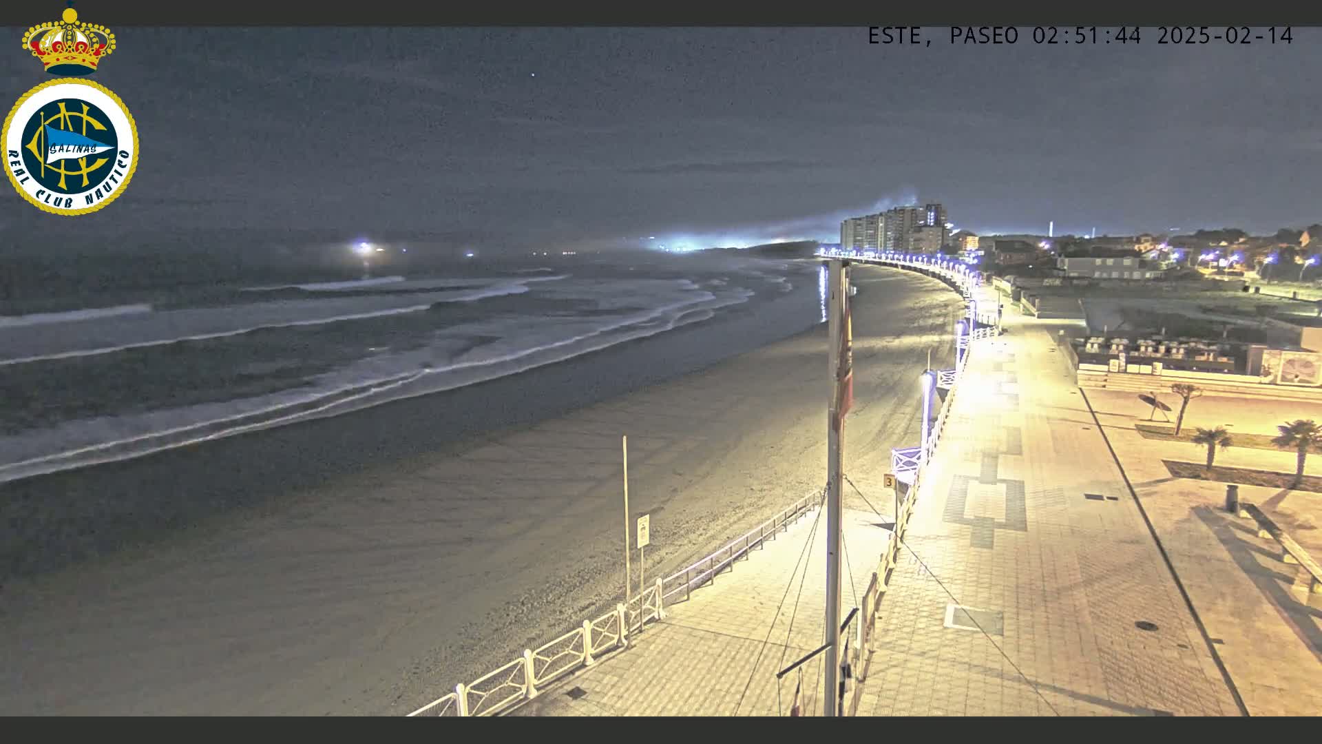 A nighttime view of a sandy beach with gentle waves under a clear, dark sky, showing a paved boardwalk lined with lights leading to buildings in the distance.