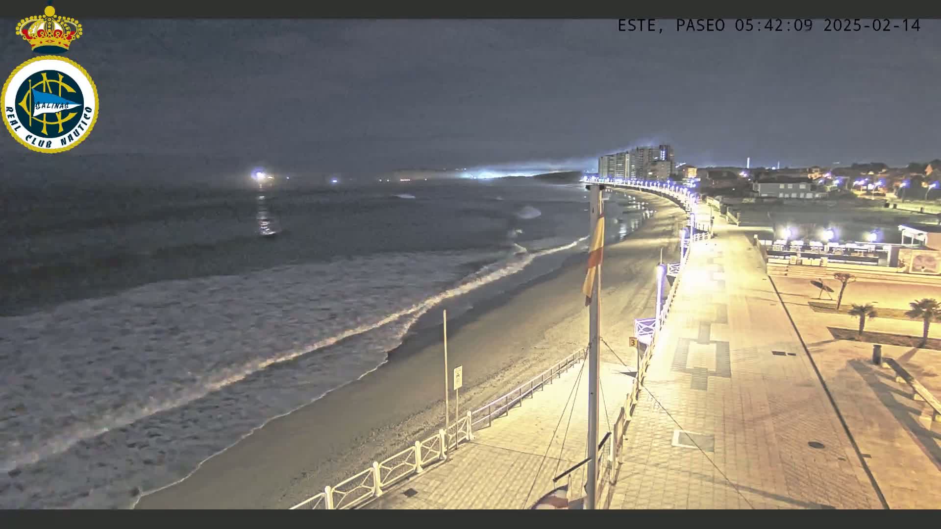 A nighttime, high-angle view shows a beach with gentle waves lapping the shore, a paved boardwalk lined with lights, and buildings in the distance.