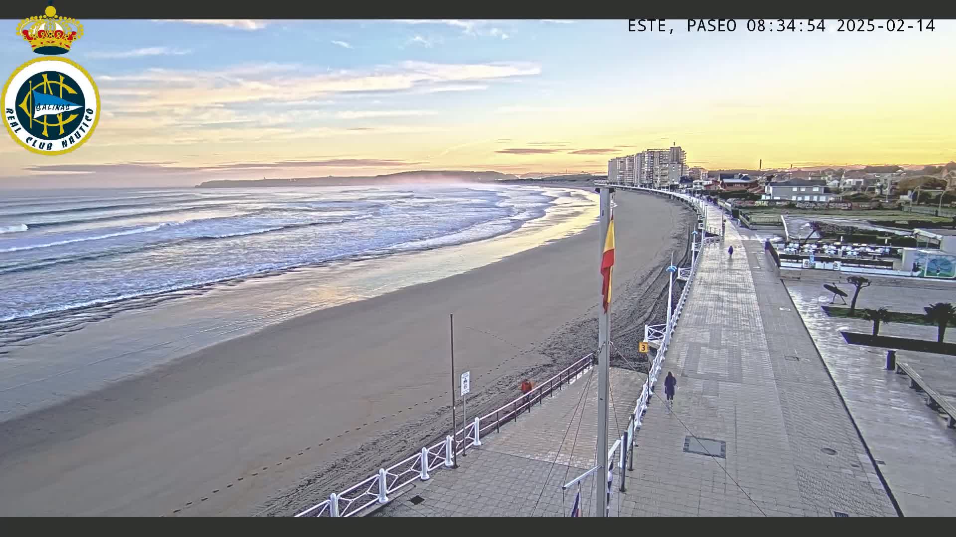 A sandy beach with gentle waves under a sunrise sky is bordered by a paved walkway and buildings.
