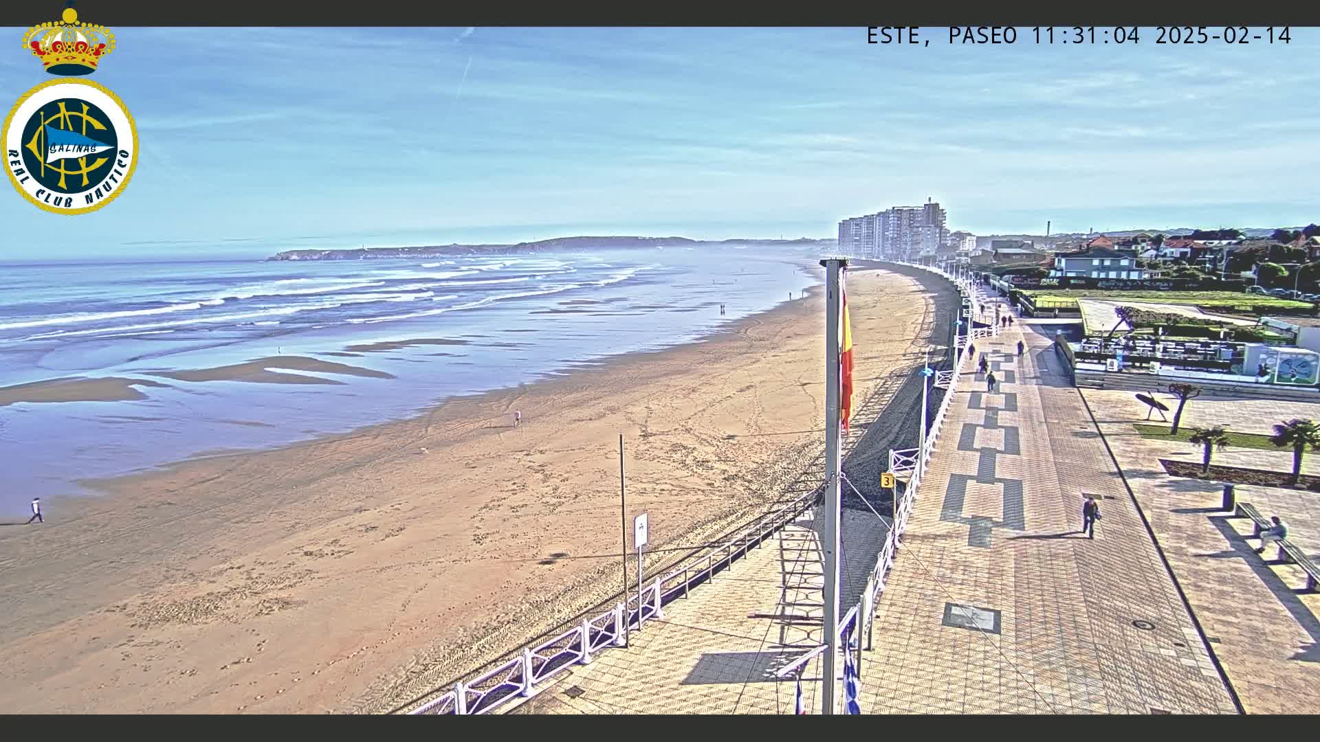 A sandy beach with gentle waves under a clear blue sky is bordered by a paved boardwalk with people strolling along it, and buildings are visible in the background.